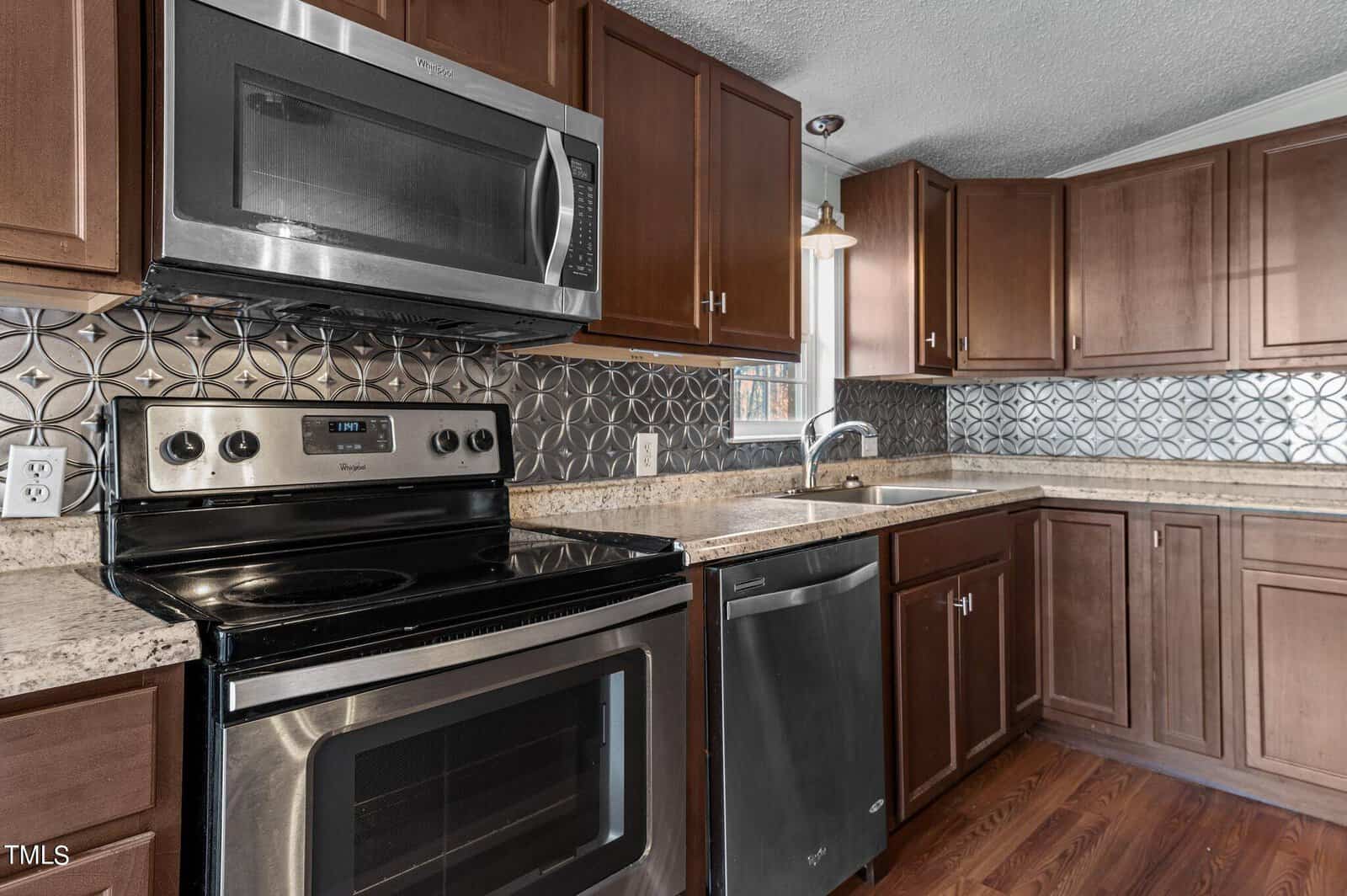 Modern kitchen with stainless steel appliances including a stove, oven, microwave, and dishwasher. Dark wood cabinets and drawers provide storage, while the countertop and patterned backsplash add an elegant touch. A window offers natural light.