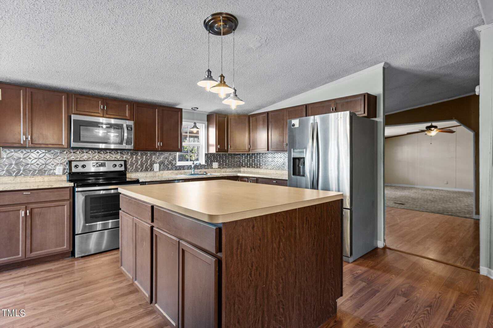 A modern kitchen with stainless steel appliances, dark wooden cabinets, and a central island with a light countertop. The backsplash features a geometric pattern. Hardwood floors extend into an adjacent room with a ceiling fan.