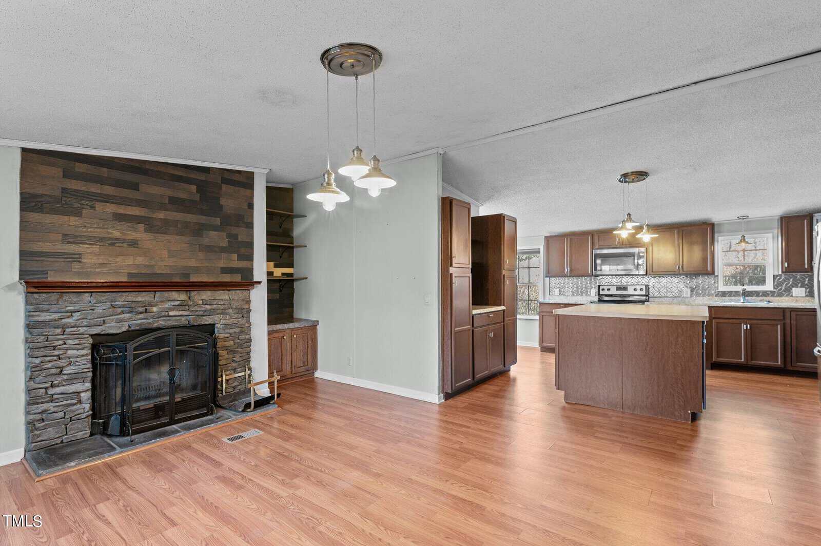 Open-plan kitchen and living room with hardwood floors. The space features a stone fireplace with a wooden mantel, modern wood cabinetry, an island, and stainless steel appliances. Pendant lights hang from the ceiling over the kitchen island.