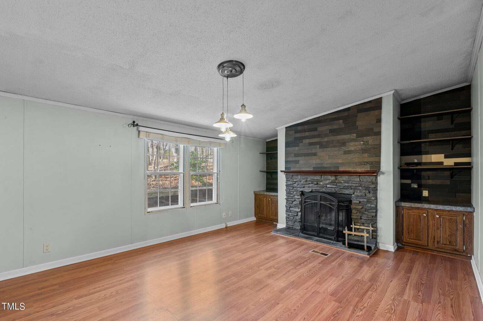 A spacious, unfurnished living room with wooden floors, a stone fireplace, and built-in shelves on both sides. A large window lets in natural light, and a ceiling light fixture hangs above. Walls are painted in a light green shade.