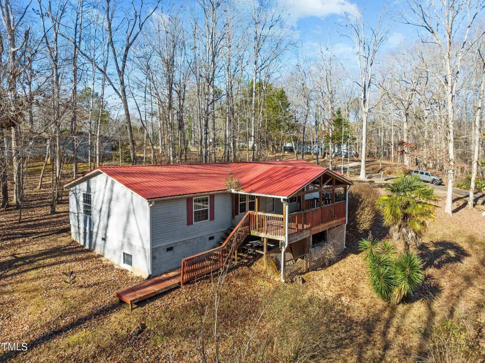 A small house with a red roof is situated in a wooded area. The home features a raised porch with wooden railings and steps leading down to the ground. Leafless trees surround the property, and a few evergreen plants are in the yard.