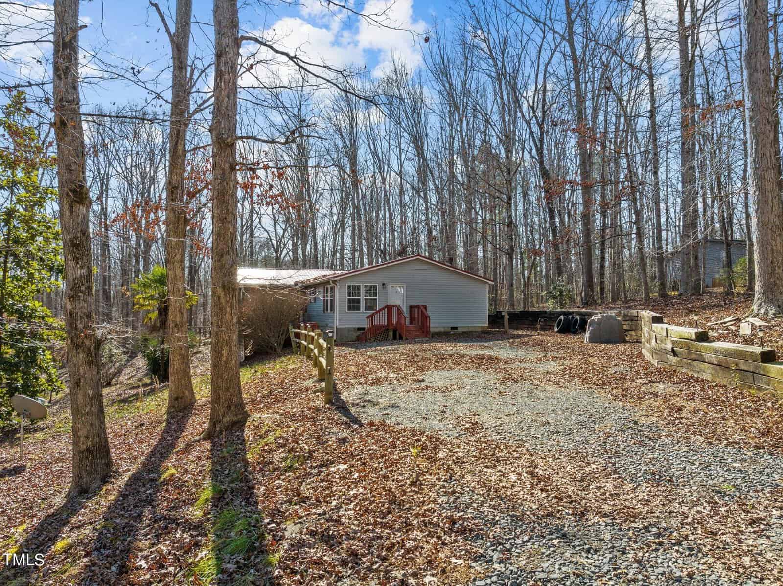 A small house with a light blue exterior and a red door sits among barren trees in a wooded area. The yard is covered with fallen leaves, and a gravel driveway leads up to the house. The sky is blue with some clouds.
