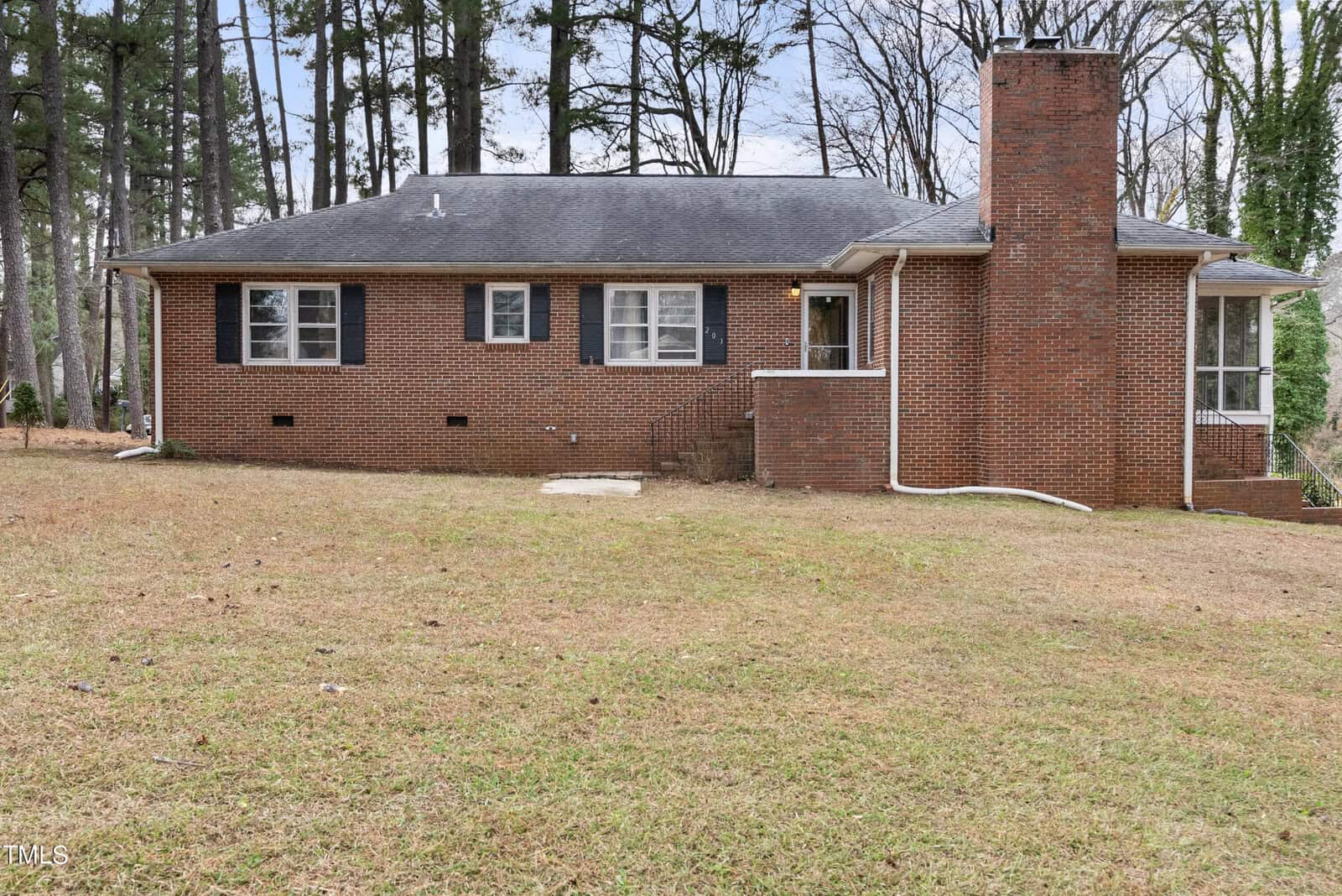 A red brick single-story house with a prominent chimney and a sloped shingled roof, surrounded by a lawn with scattered trees, under a cloudy sky.