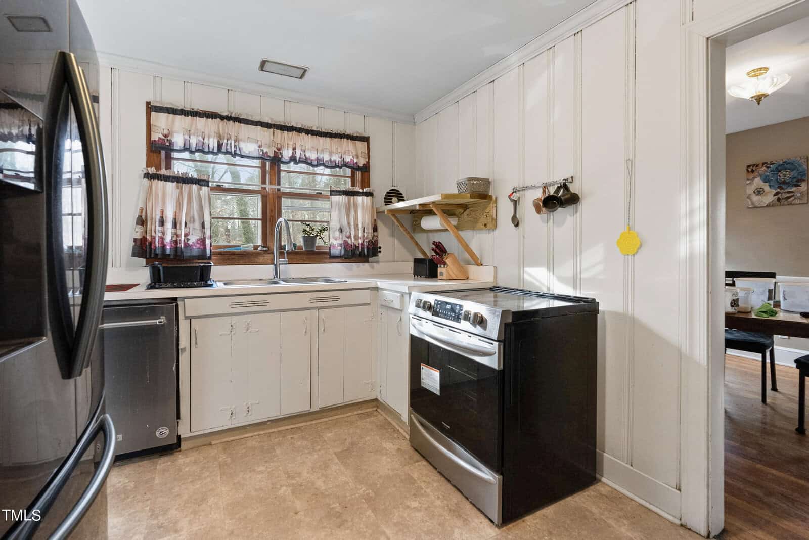 A modestly-sized kitchen with white paneled walls, featuring a stainless steel stove and refrigerator, with a window above the sink that has patterned curtains. a wooden shelf holds a few items on the wall to the right of the window.