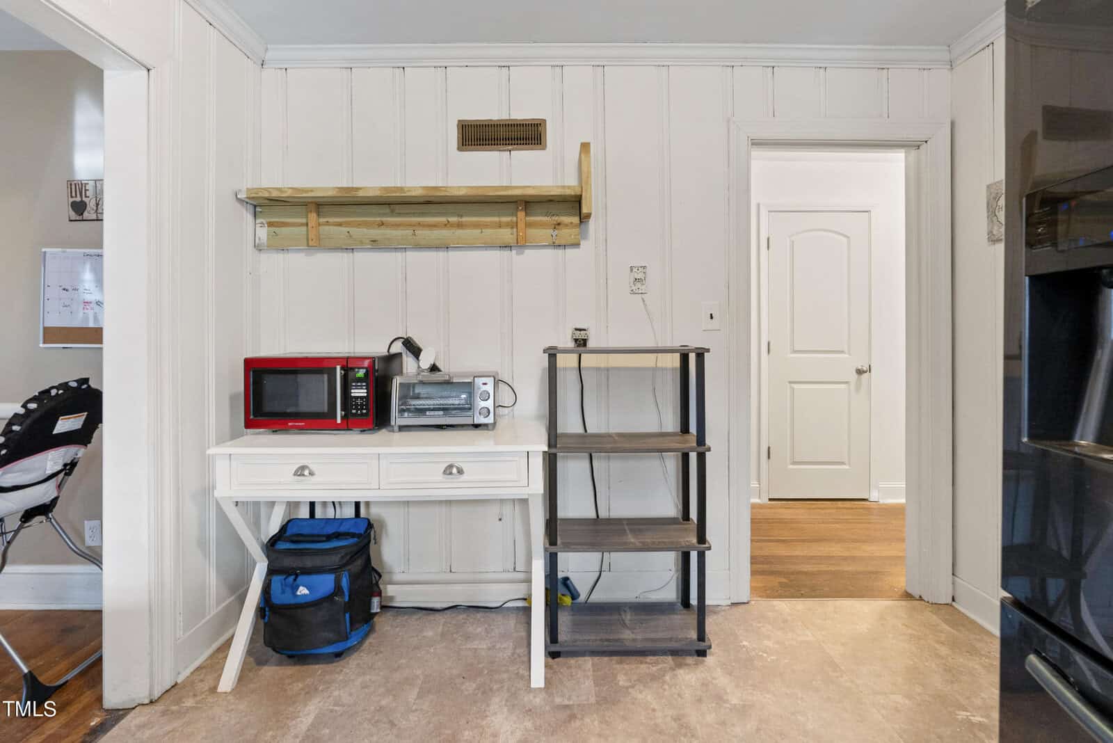 A compact kitchen nook with a white desk, shelving units, a microwave, and a coffee maker, adjacent to a hallway with an ajar door.