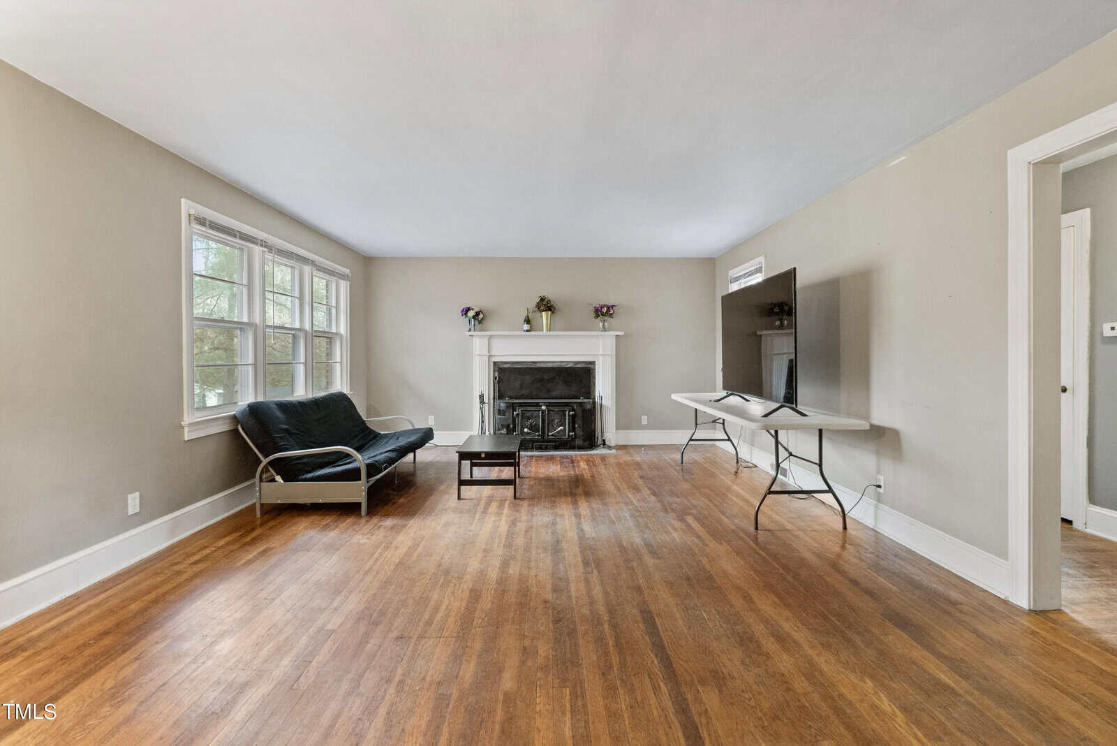 A spacious living room with hardwood floors, featuring a black armchair, a coffee table, a portable table, and a fireplace with a white mantel. there is a window on the left providing natural light, and the walls are painted in a light neutral color.