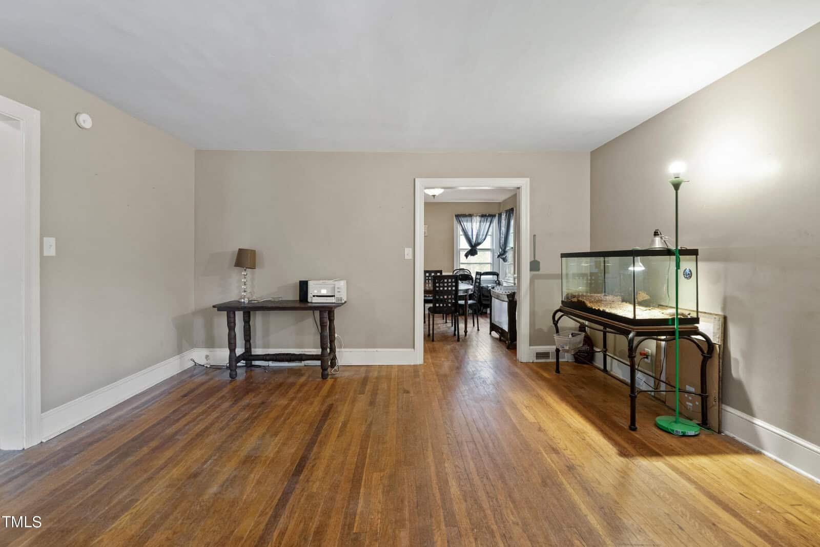 A spacious room with hardwood flooring featuring a small desk with a laptop, a standing lamp, and a glass terrarium on a metal stand. the room opens up to another area with a dining set visible in the background.