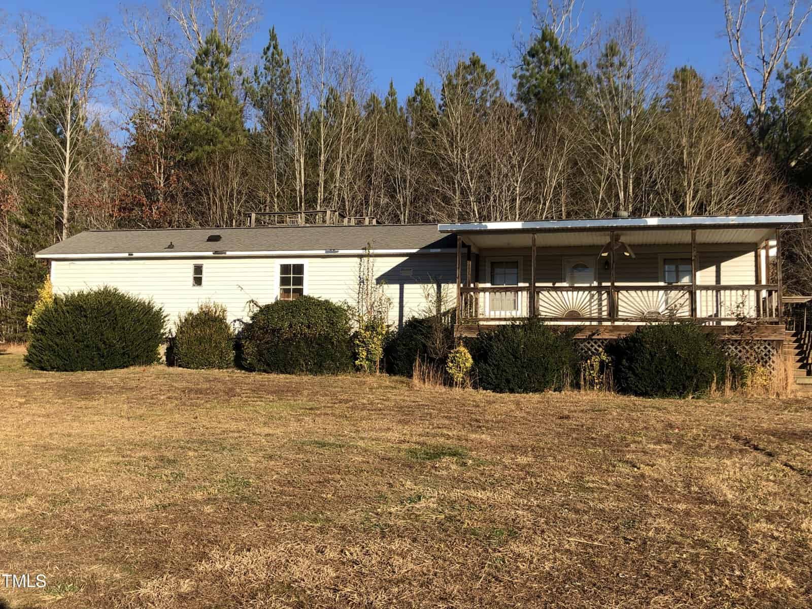 Nestled on 8178 Raymond Royster Road in Roxboro, this single-story house features white siding and a charming wraparound porch, beautifully set against a backdrop of tall trees. The grassy front yard is dotted with bushes, all under a clear blue sky.