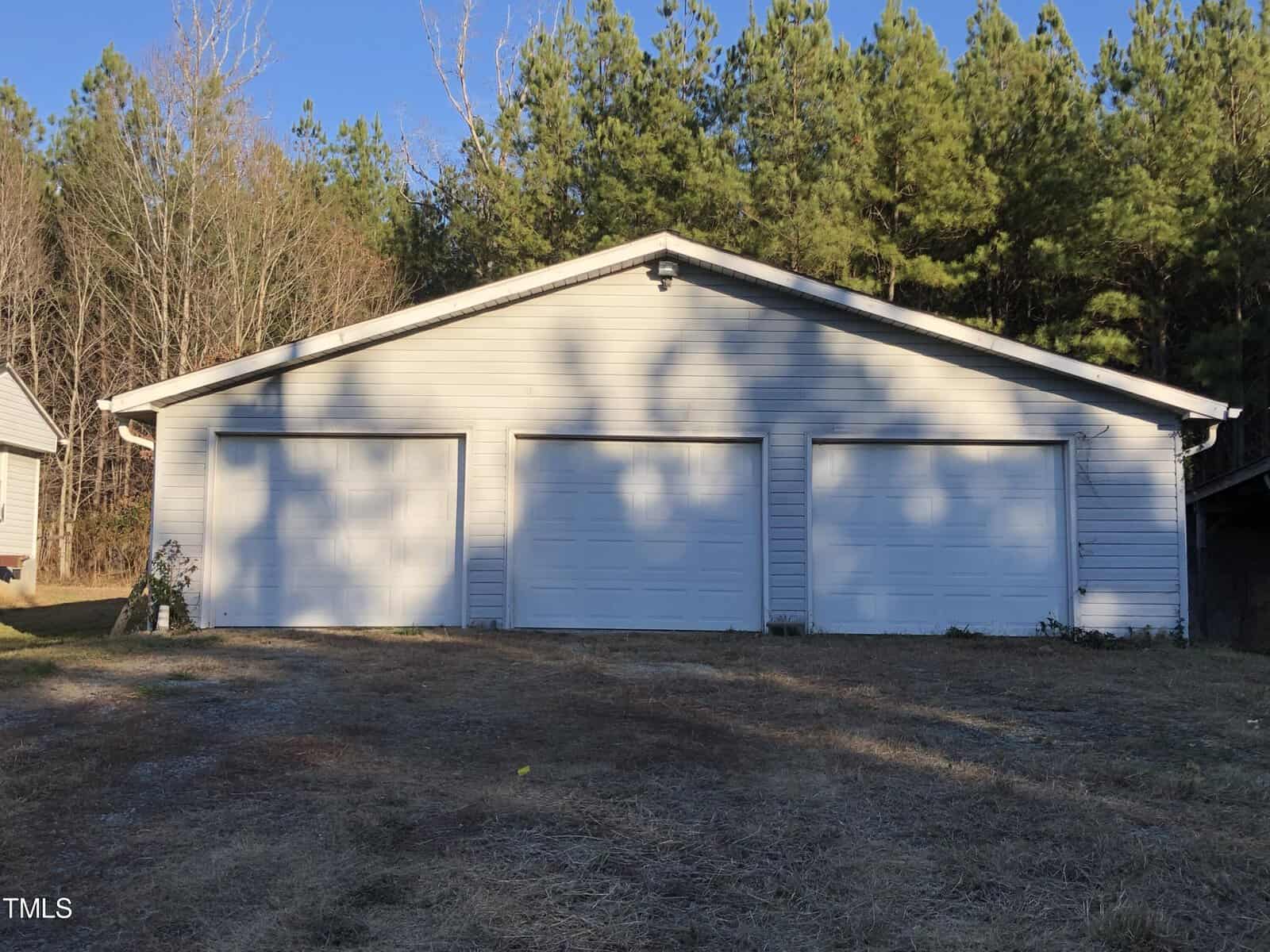 A white garage with three closed doors is situated in a rural area, surrounded by trees. The ground in front appears to be bare and grassy, with visible patches of sunlight and shadow cast by the trees.