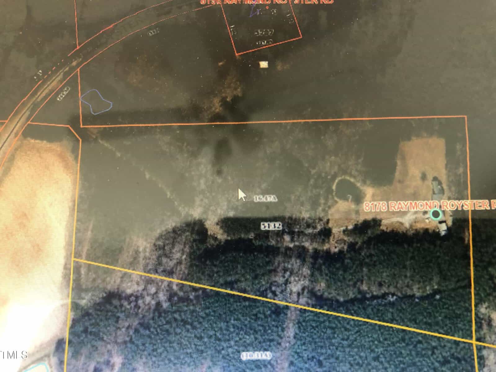 Aerial view of land parcels outlined in orange and yellow lines. Most of the area is covered with dense green foliage, with a few clearings. A road curves at the top left corner, and text indicates parcel numbers and a name: "3178 Raymond Royster.