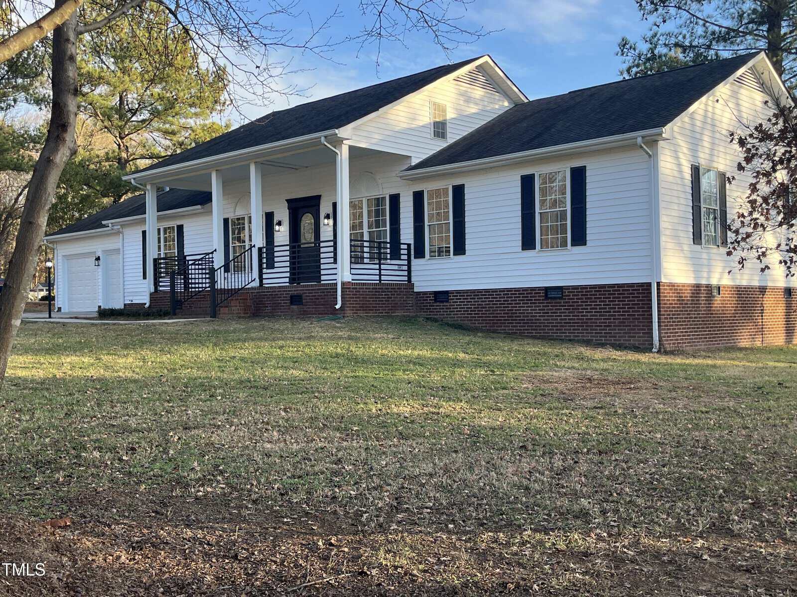 A single-story house with white siding, a brick foundation, and black shutters, featuring a front porch with stairs and railings, surrounded by a grassy lawn with several trees in the background.