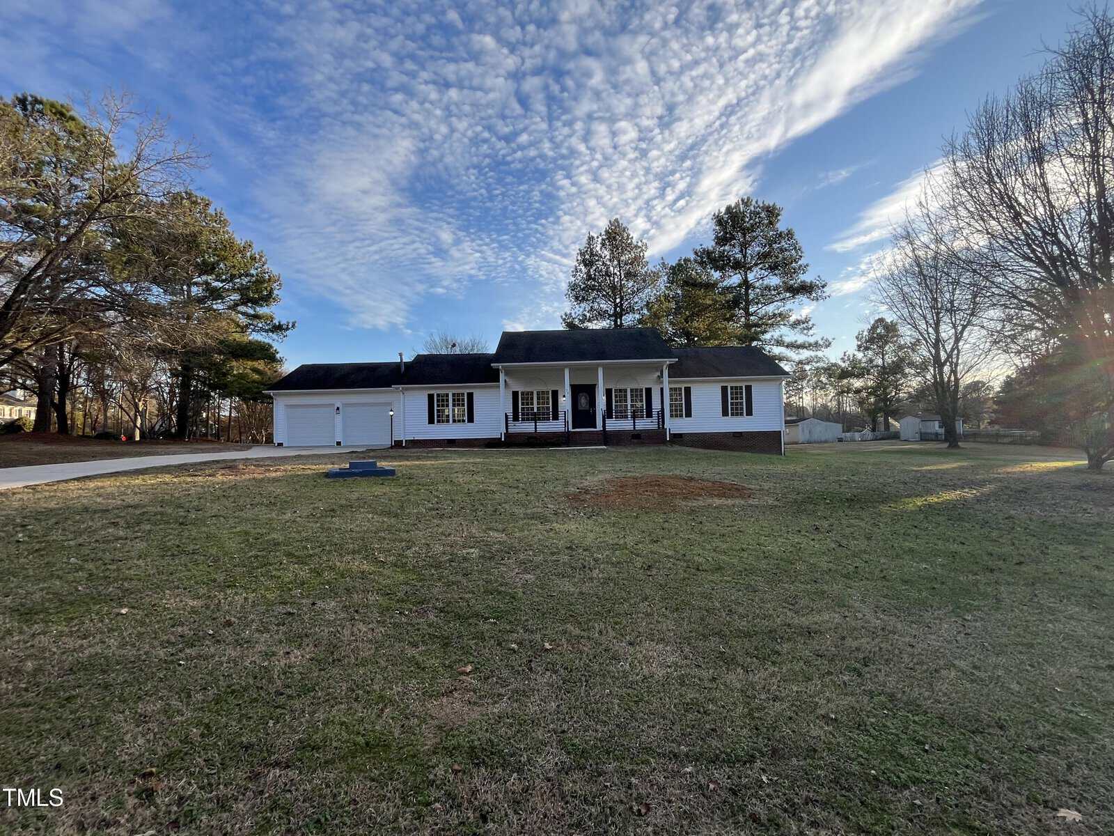 A single-story house with a front porch situated in a grassy area with trees in the background under a cloudy blue sky.