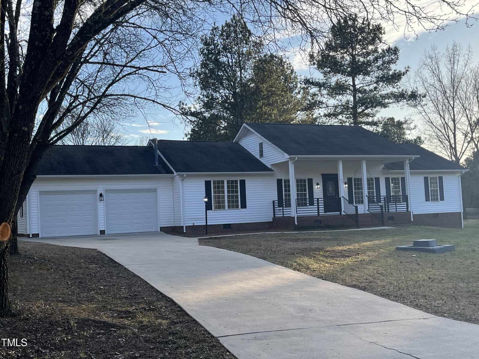 Single-story residential home with white siding and a black shingle roof, featuring two garage doors and a covered front porch, set against a backdrop of trees with a driveway leading up to the garage.