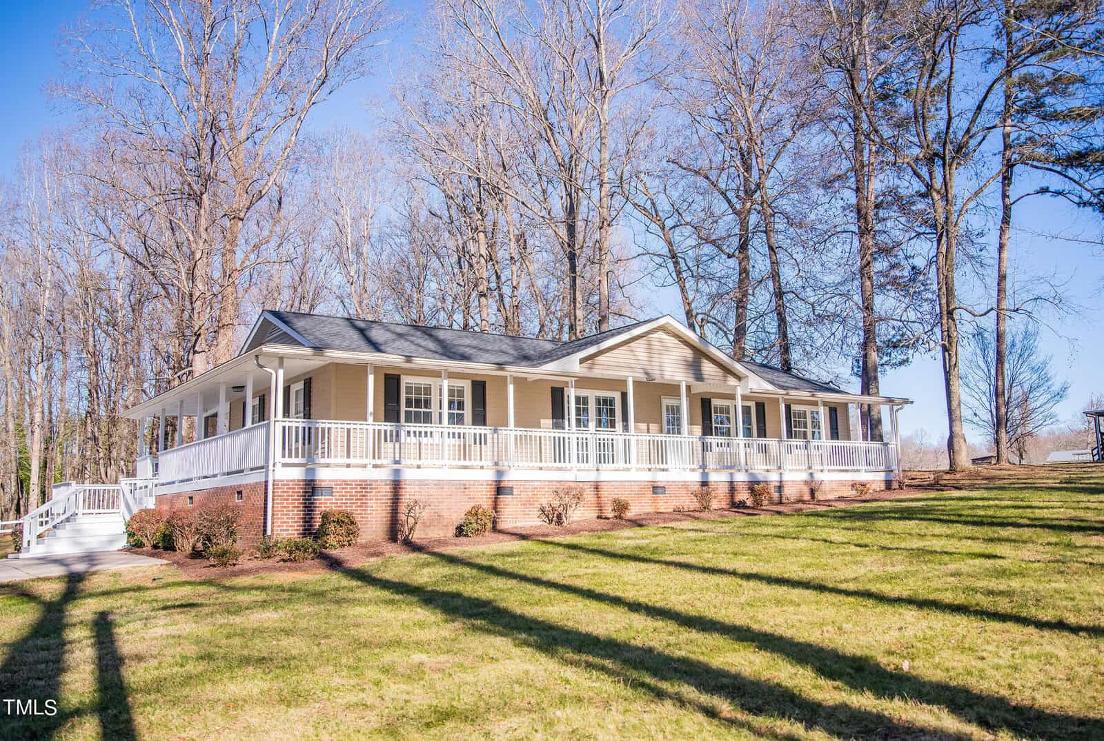 A ranch-style house with light brown siding and a large wrap-around porch, set amidst tall trees and a freshly cut lawn. The house features a pitched roof and brick foundation, with bare trees indicating a winter or early spring setting.