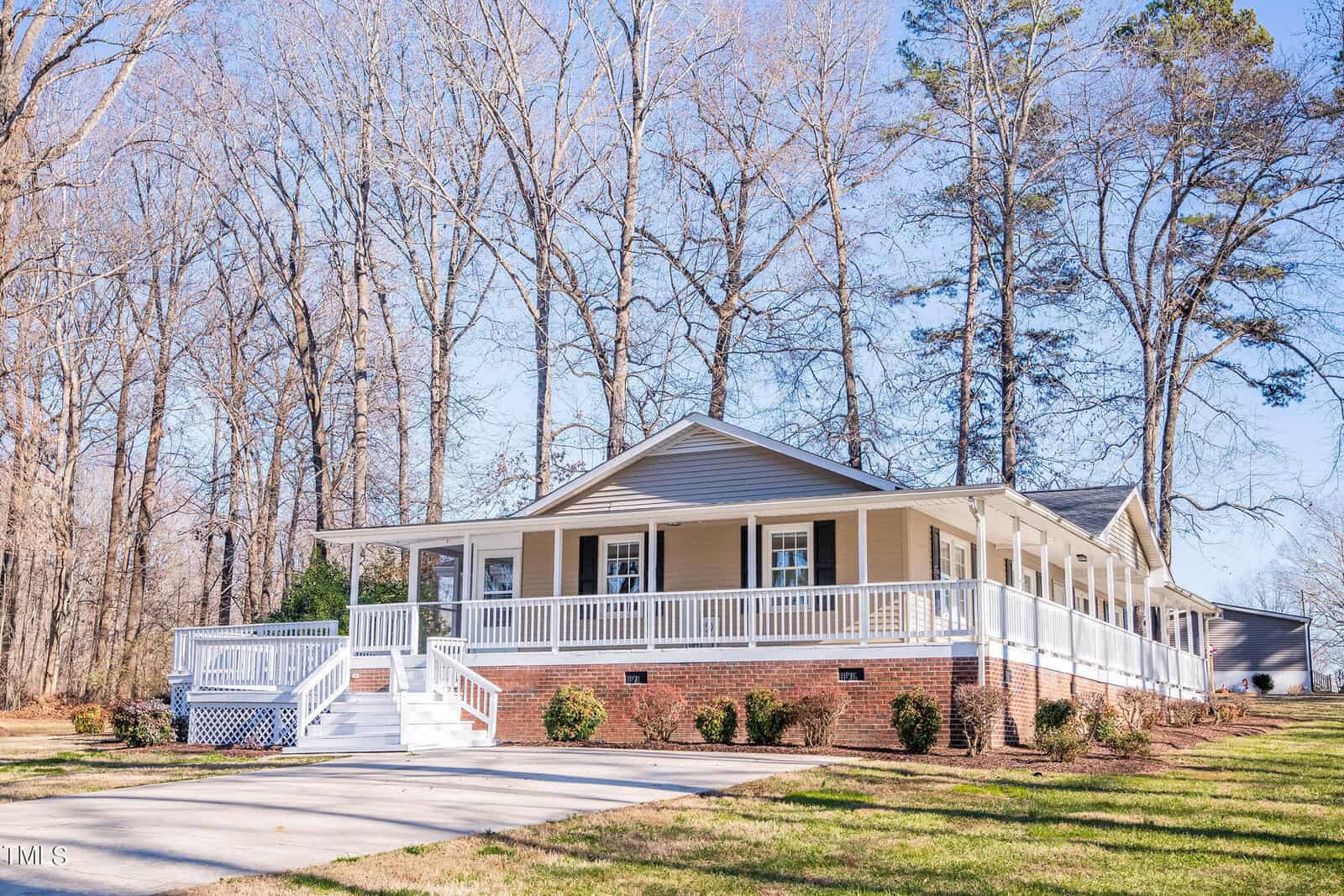 A single-story house with a wraparound porch surrounded by bare trees. The house features tan siding, a brick foundation, and white railings. The yard is spacious with a concrete driveway leading to the porch steps.