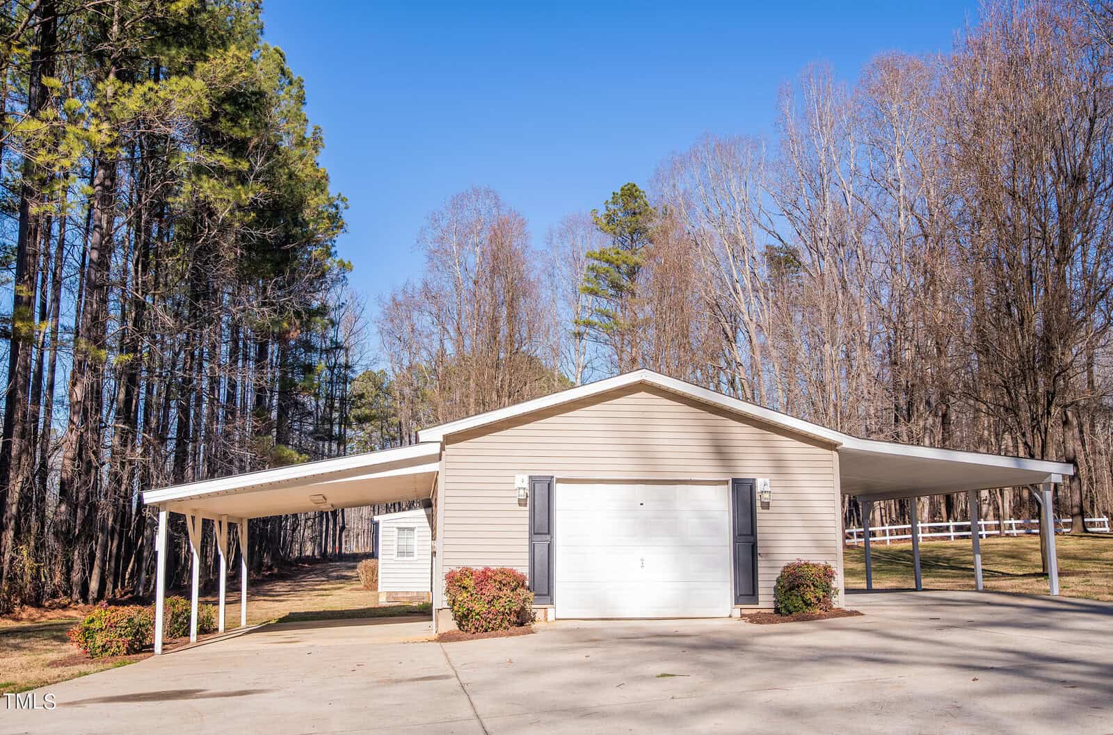 A single-story beige house with a white garage door, surrounded by tall trees. The home features two carports on either side, and a large paved driveway leading up to it under a clear blue sky.