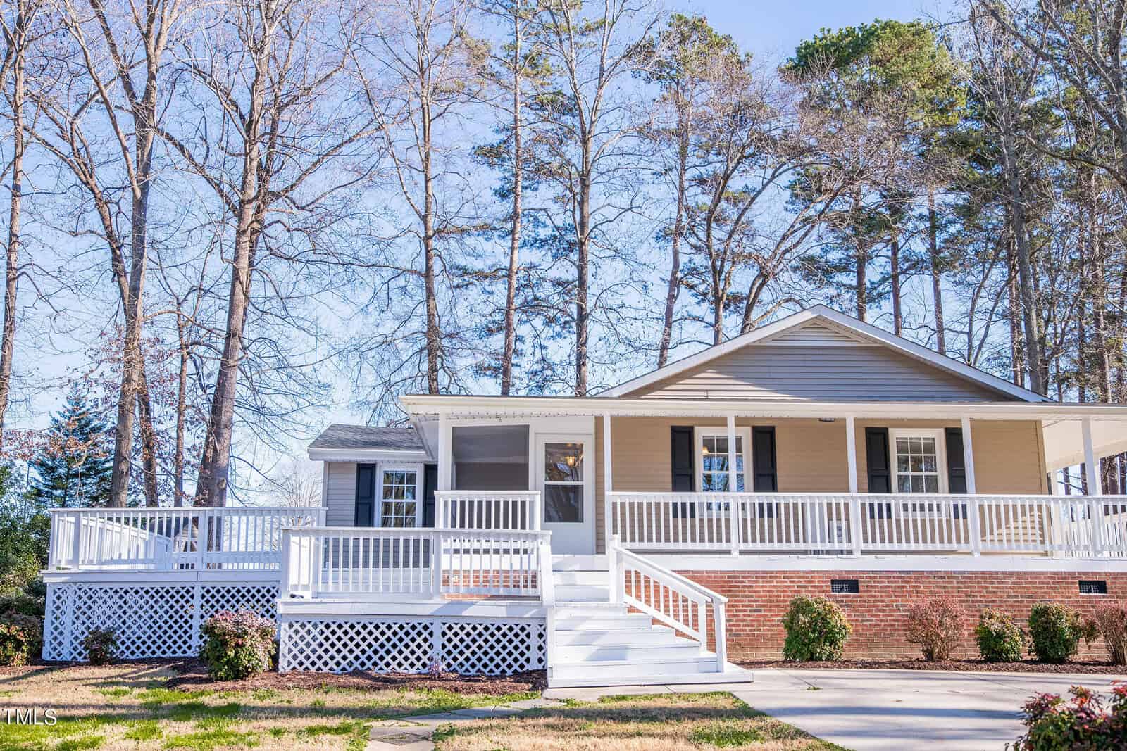 A charming single-story house with beige siding and a wide front porch, surrounded by tall trees. The porch features white railings and steps leading to the front door. The front yard is landscaped with shrubs.