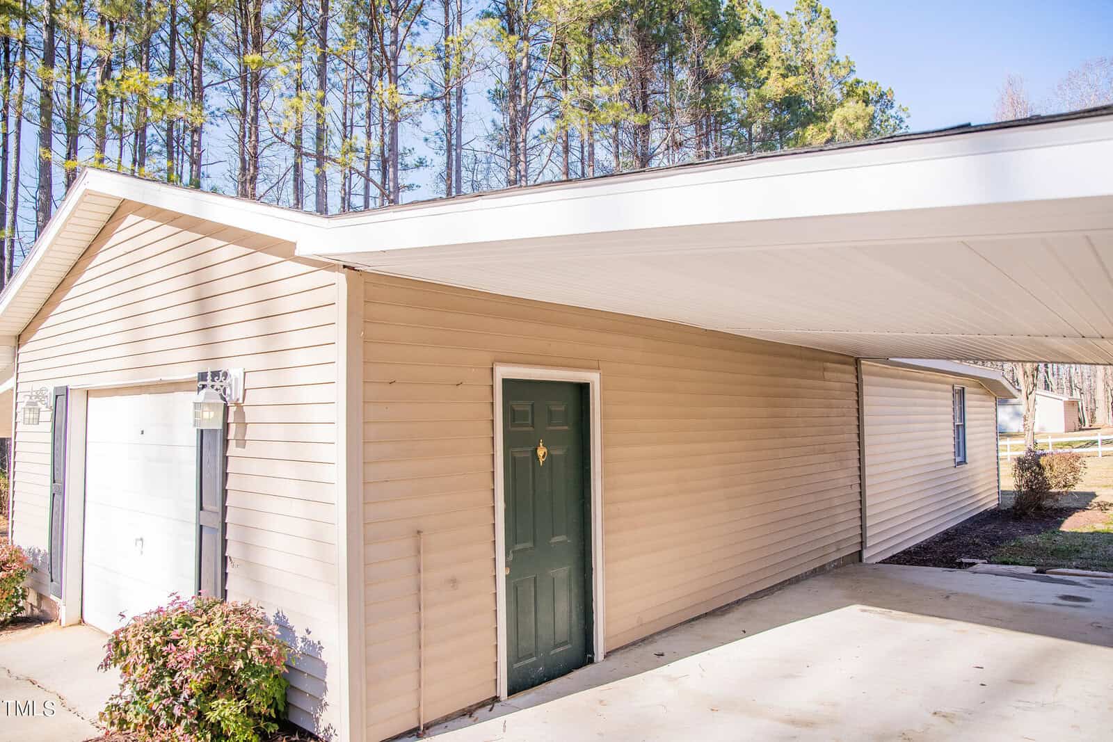 A beige garage and carport are shown, with a green door on the side of the garage. The carport is attached to the garage, and the area is surrounded by tall pine trees under a clear blue sky. Shrubs accent the garage exterior.