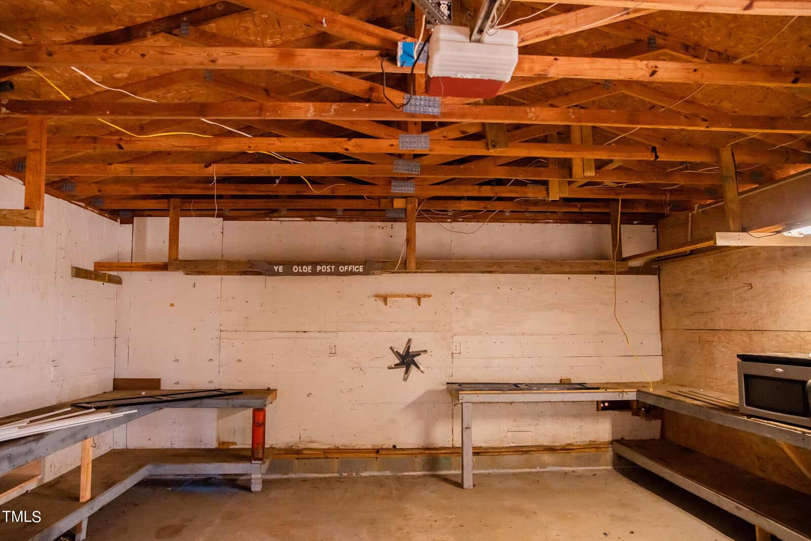 Interior of a wooden workshop with a slanted ceiling and exposed beams. The room features long workbenches against white walls, with a star-shaped decor hanging. A small sign reads "Ye Olde Post Office." A microwave is on the right.