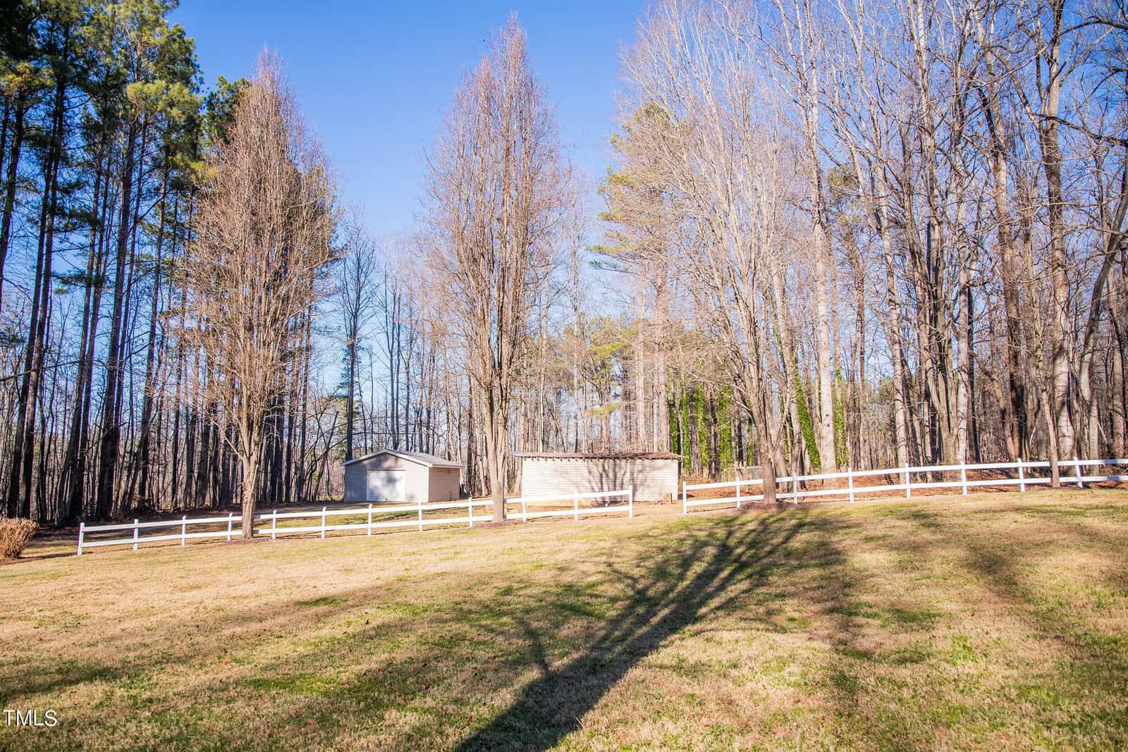 A serene landscape with a grassy lawn, bordered by bare trees under a clear blue sky. A small white fence encloses a simple structure, possibly a shed, adding rustic charm to the natural setting. Sunlight casts long shadows on the grass.