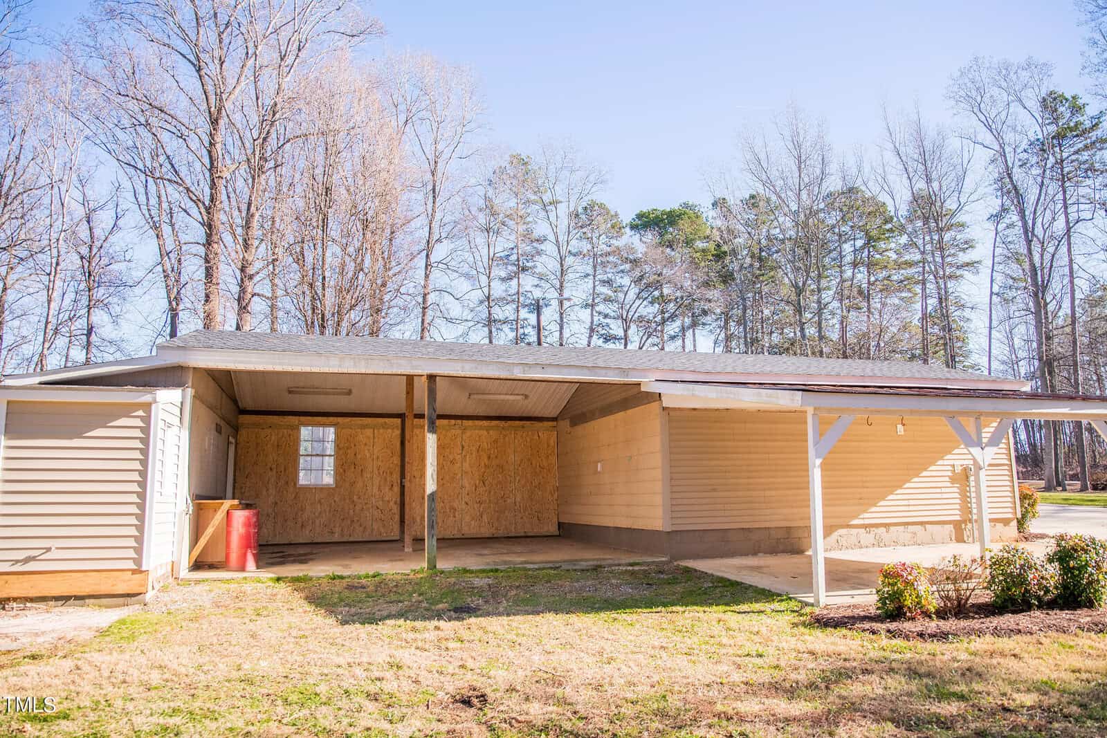 A small, single-story wooden building with a covered carport attached on the right side. The surrounding area has grass and some shrubs, and leafless trees can be seen in the background against a clear sky.