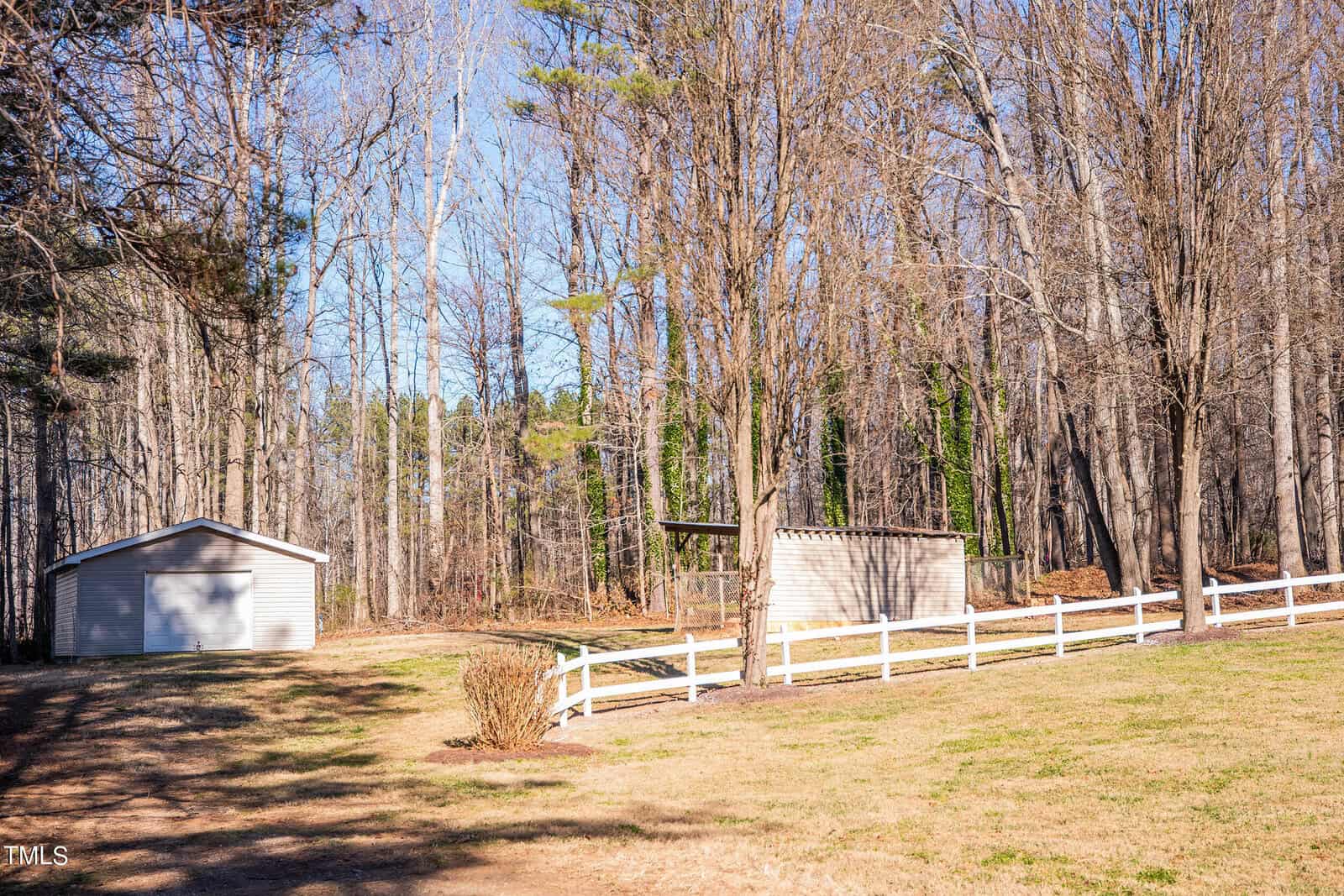A rural scene featuring a small shed and a fenced area surrounded by bare trees. The sky is clear blue, and the ground is a mix of grass and sparse vegetation. A larger building is also visible on the left.