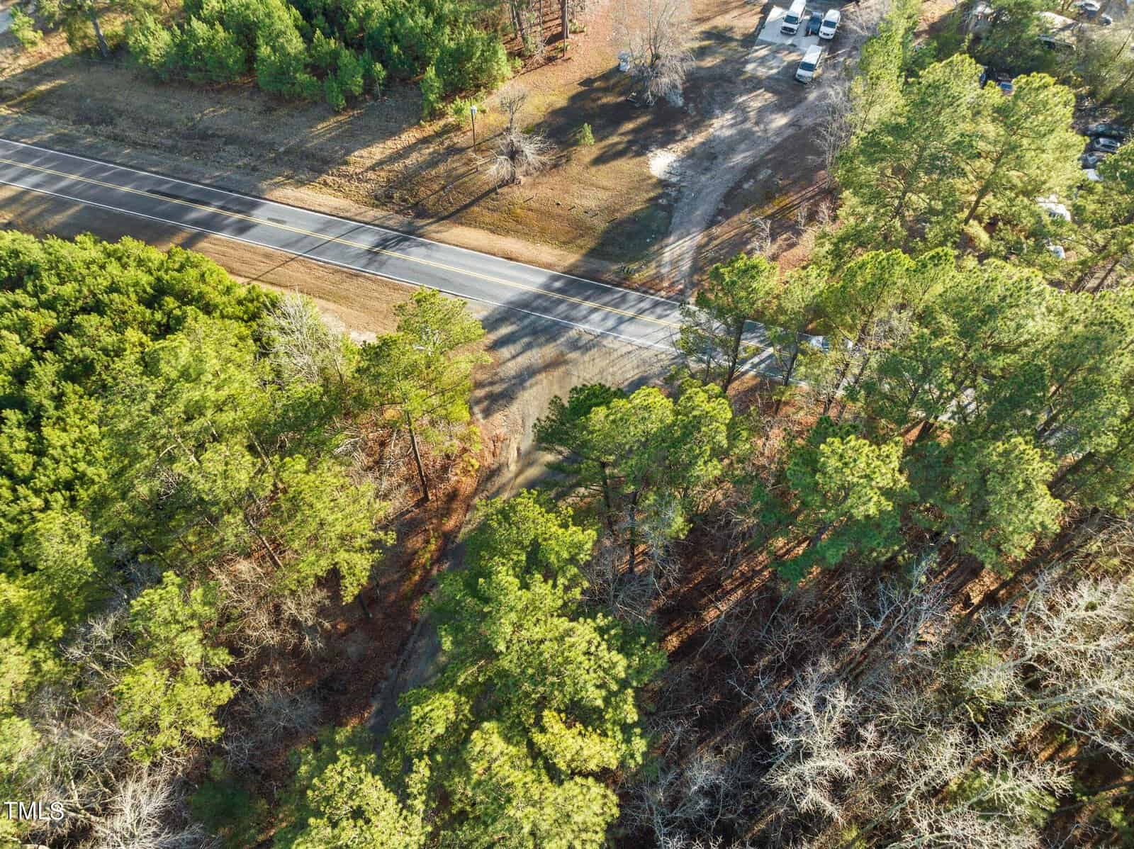 Aerial view of a forested area with a road cutting through it diagonally. Several cars are parked on a dirt lot near the road. The trees are a mix of evergreen and deciduous, with some areas showing bare branches.