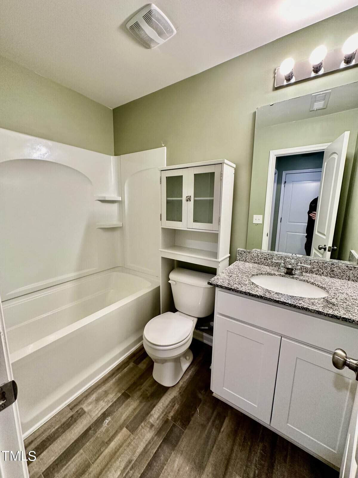 A modern bathroom featuring a white bathtub-shower combo, toilet, grey vanity with a granite countertop, and wood-effect flooring, with a reflection of a person in the mirror.
