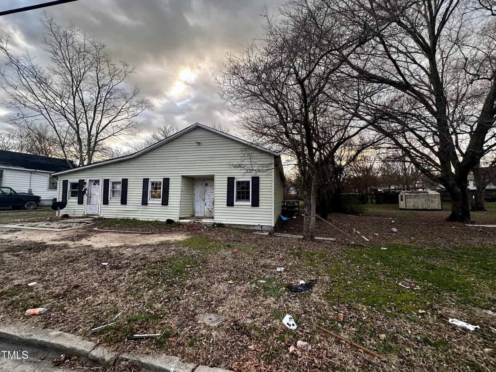 A single-story, white house with boarded-up windows and a littered front yard under a cloudy sky.