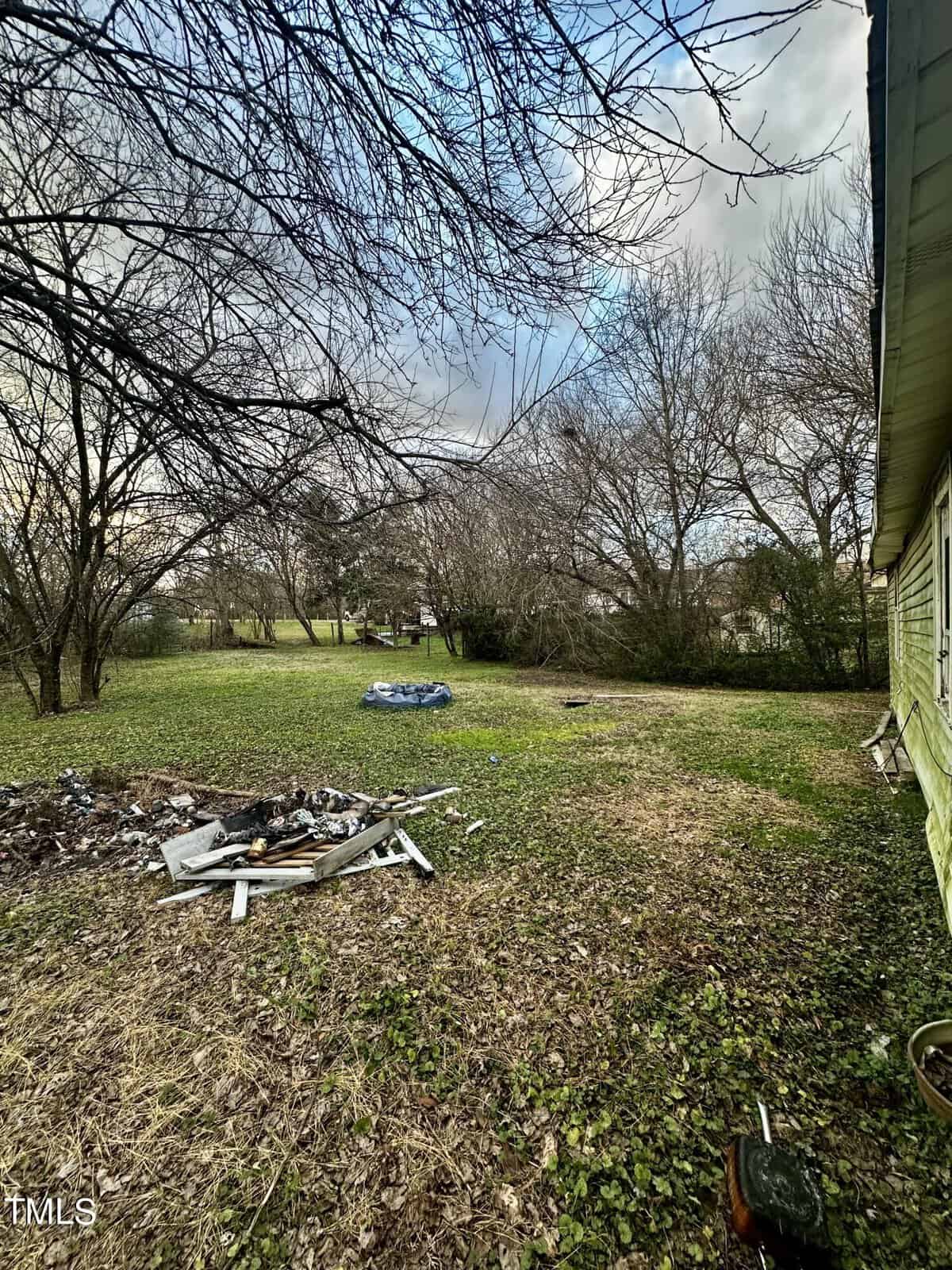 A backyard with scattered debris and leafless trees under a cloudy sky, with a tarp-covered object in the distance.