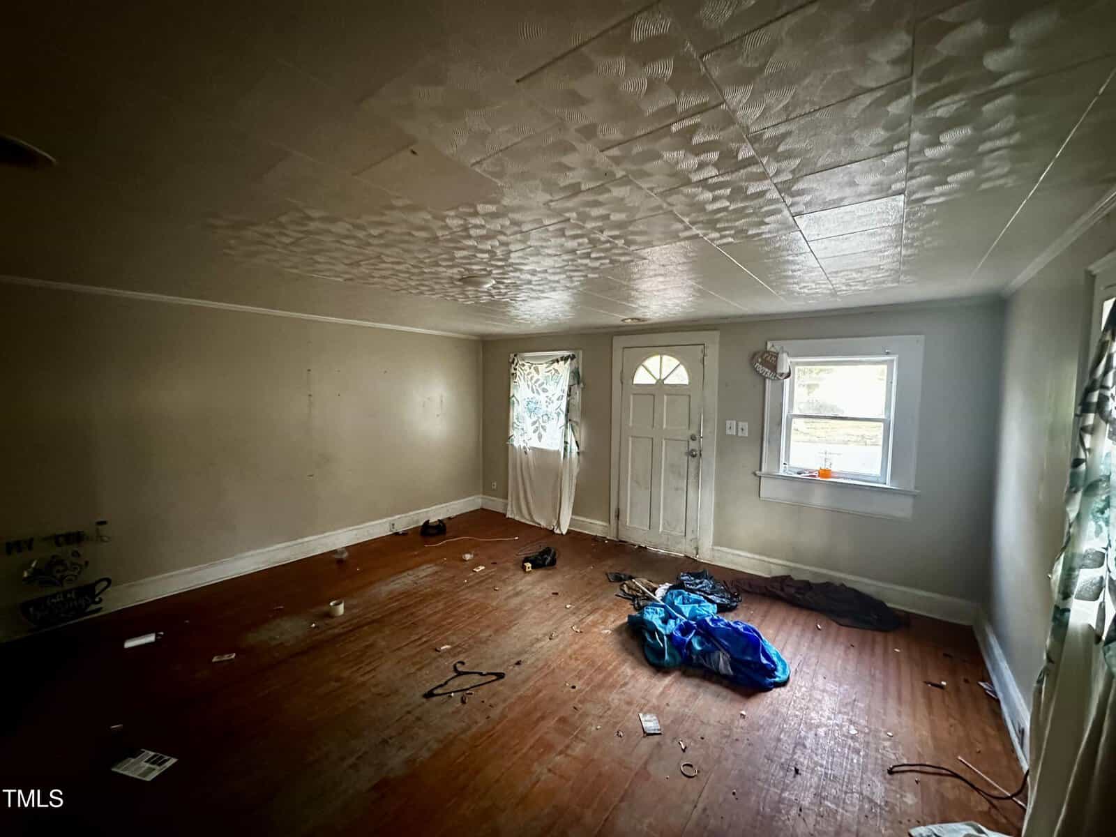 An empty room with a textured ceiling and hardwood flooring, showing signs of wear and some debris on the floor. the room has a closed white front door with decorative glass and a window allowing in natural light.
