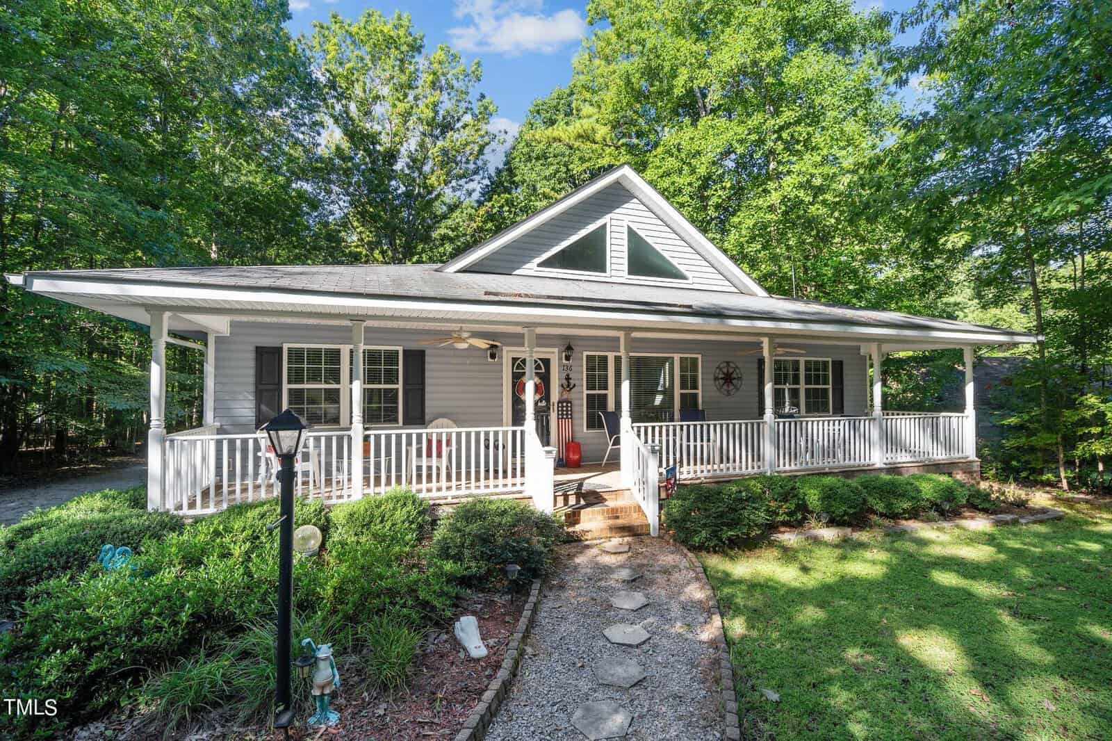 A single-story house with a large front porch surrounded by trees on a sunny day.