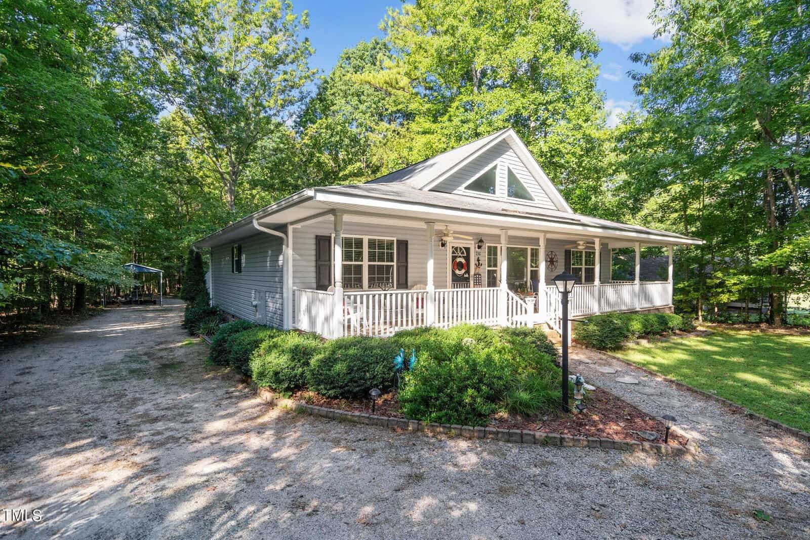 A single-story white house with a gabled roof, surrounded by trees, featuring a small front porch and a gravel driveway.
