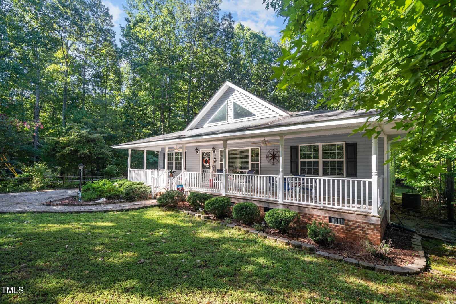A single-story house with white siding and a front porch, nestled among trees with a cleared lawn area and a driveway to the left.