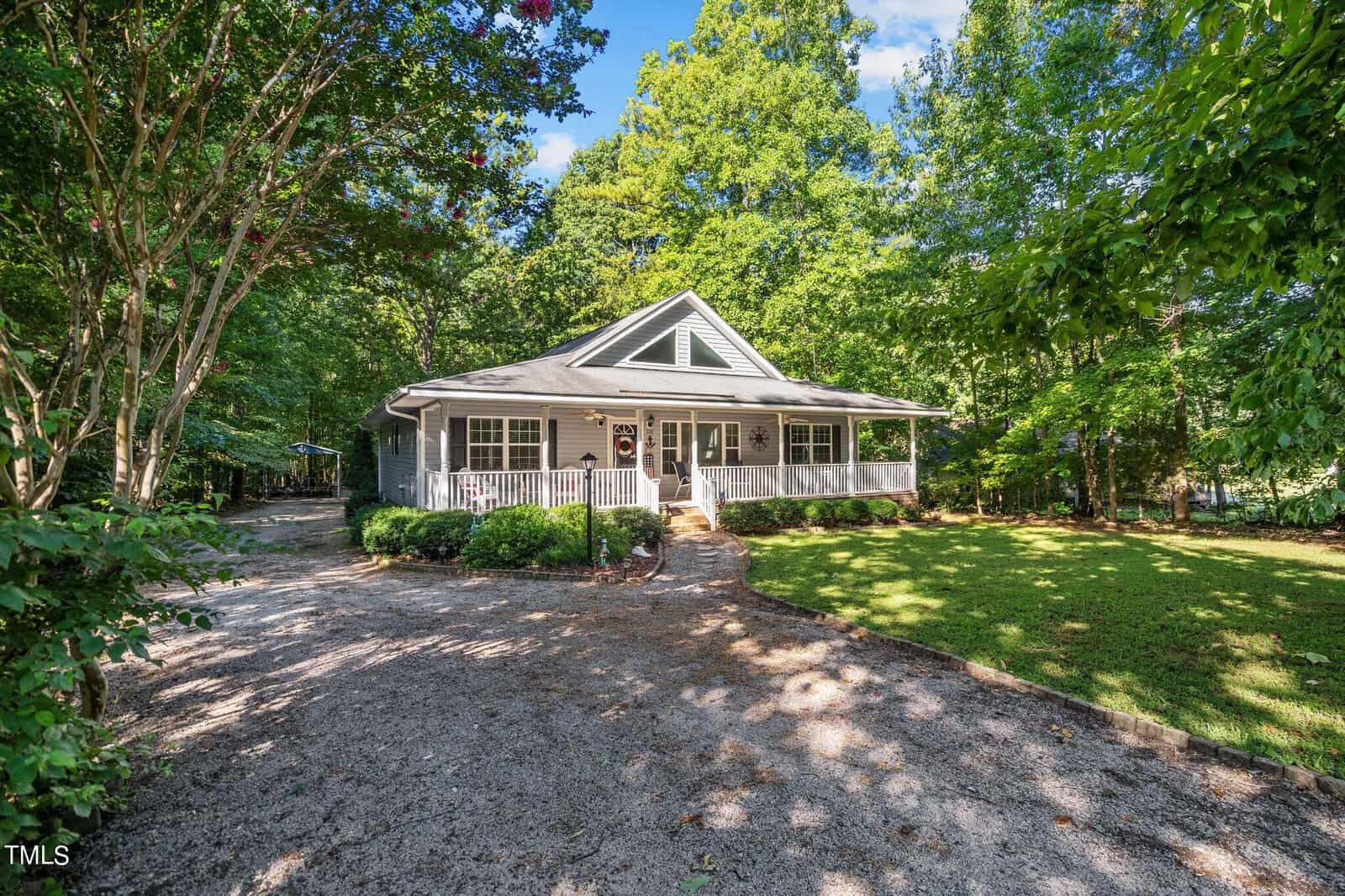 A single-story house with a gabled roof and a covered front porch, surrounded by lush greenery and a gravel driveway.