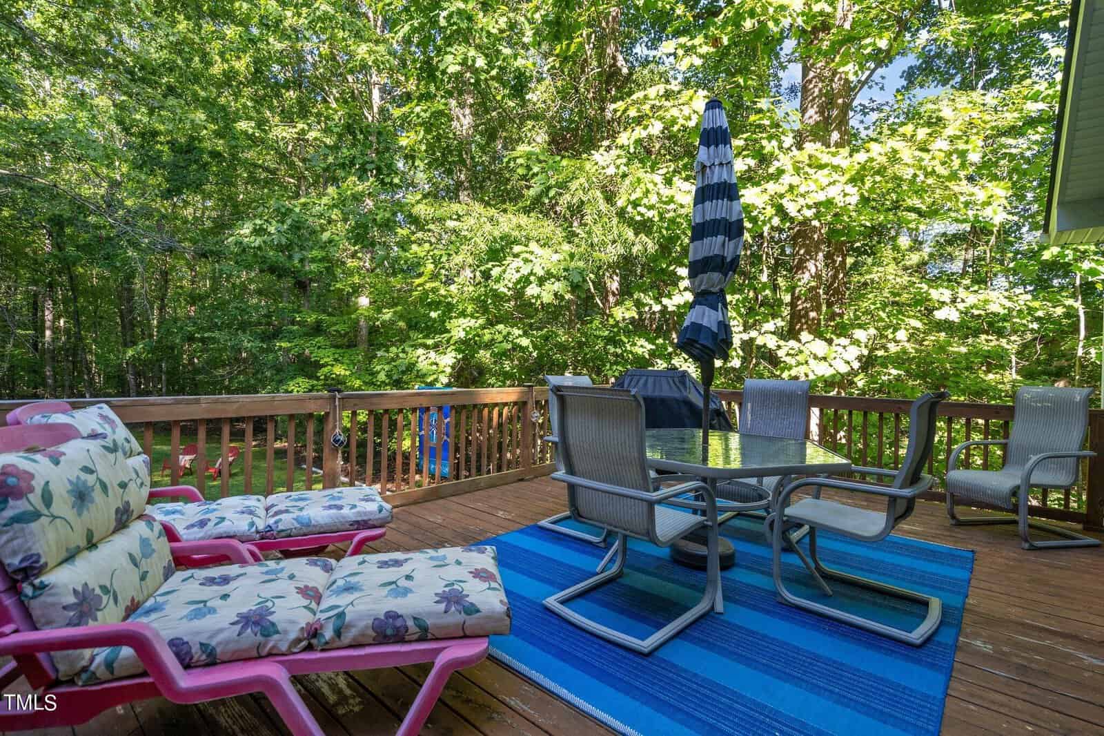 A sunny day on a residential deck surrounded by trees, featuring outdoor furniture with a closed umbrella, a sun lounger, and a patterned blue rug.