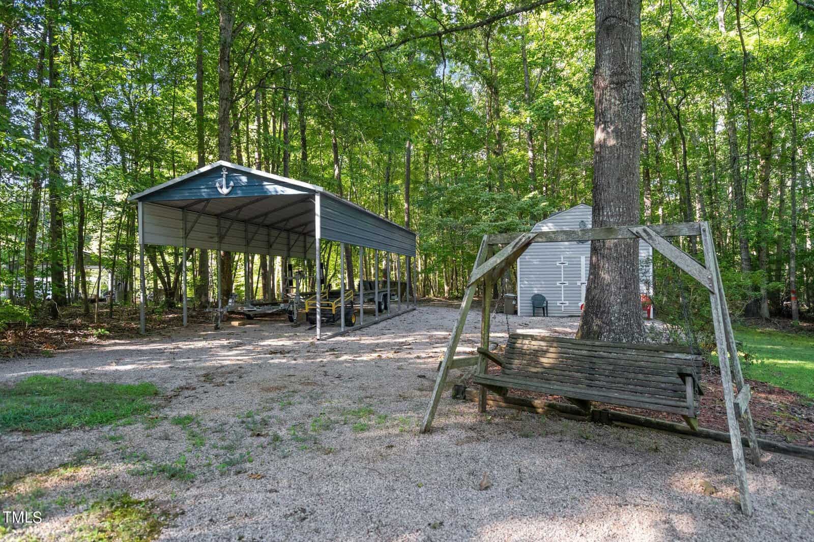 A wooden swing sits in a gravel-covered yard surrounded by tall trees, with a metal carport to the left and a small gray storage shed to the right.