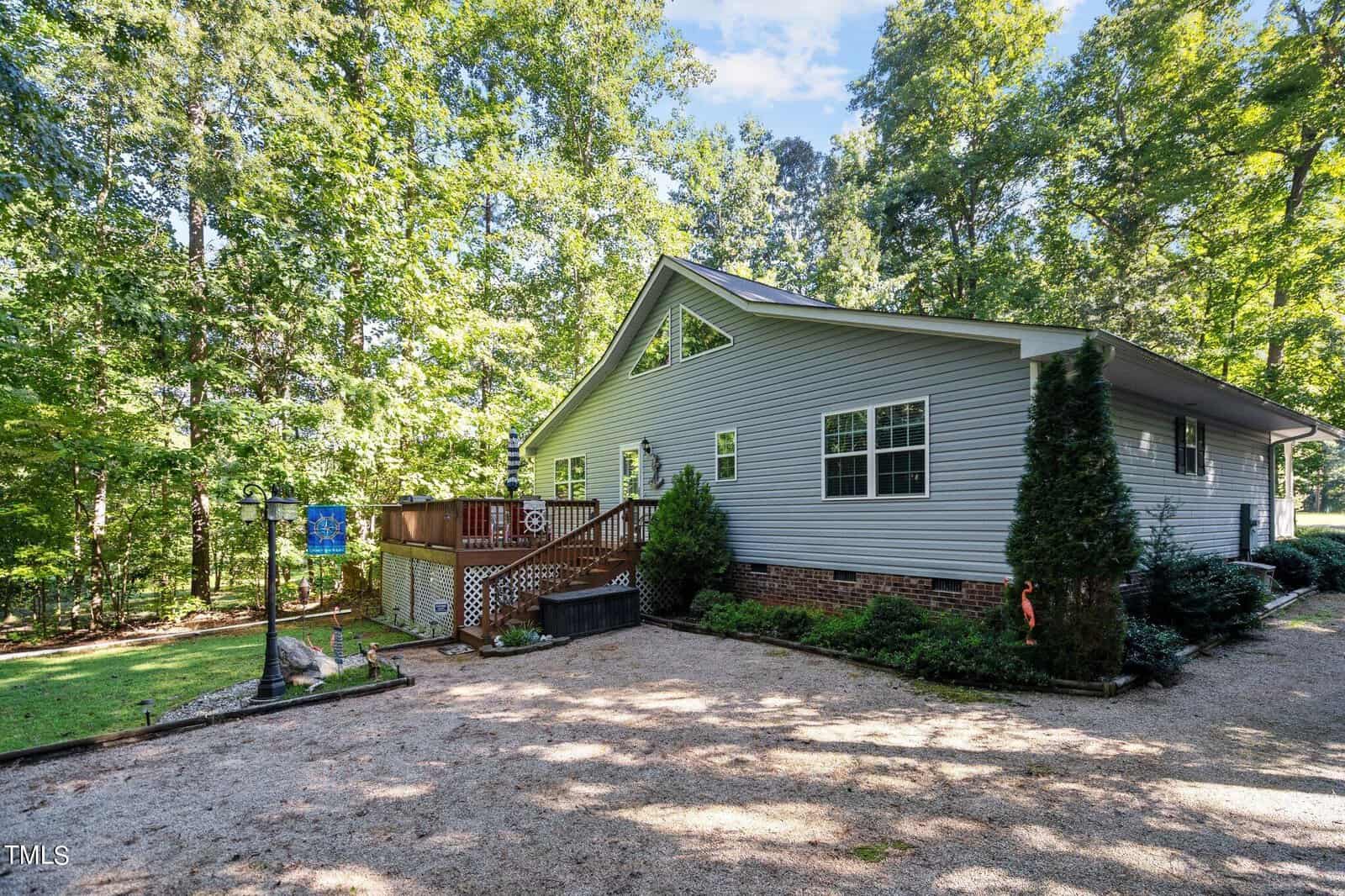 A single-story house with light blue siding, surrounded by trees, featuring a small deck and a driveway on the side.