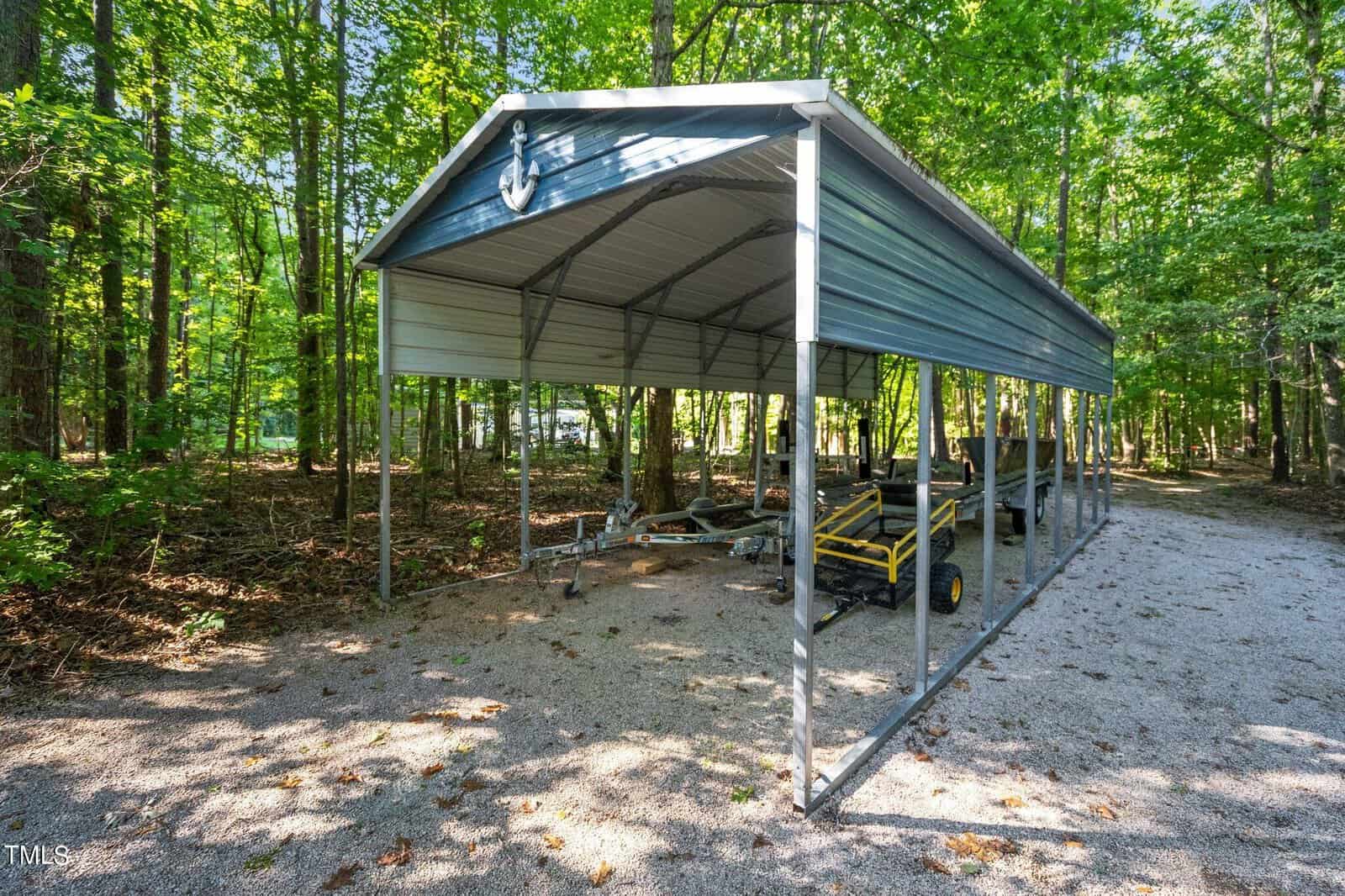 A metal carport shelter in a wooded area, housing a yellow utility vehicle and various equipment.