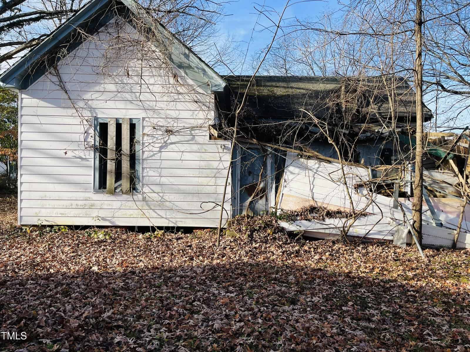 A weathered, rundown house with peeling white paint and a sagging roof. Windows are broken, and leafless trees surround the structure. The ground is covered in dry leaves, indicating an autumn season.