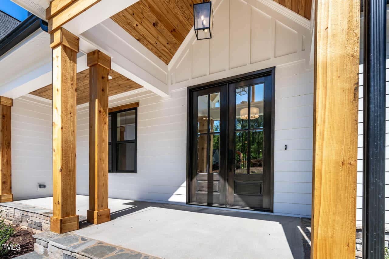 Modern house porch with wooden beams, white siding, and black-framed glass doors. The ceiling features warm-toned wood paneling and a hanging light fixture. A stone pathway leads to the entrance, surrounded by a green lawn in the background.