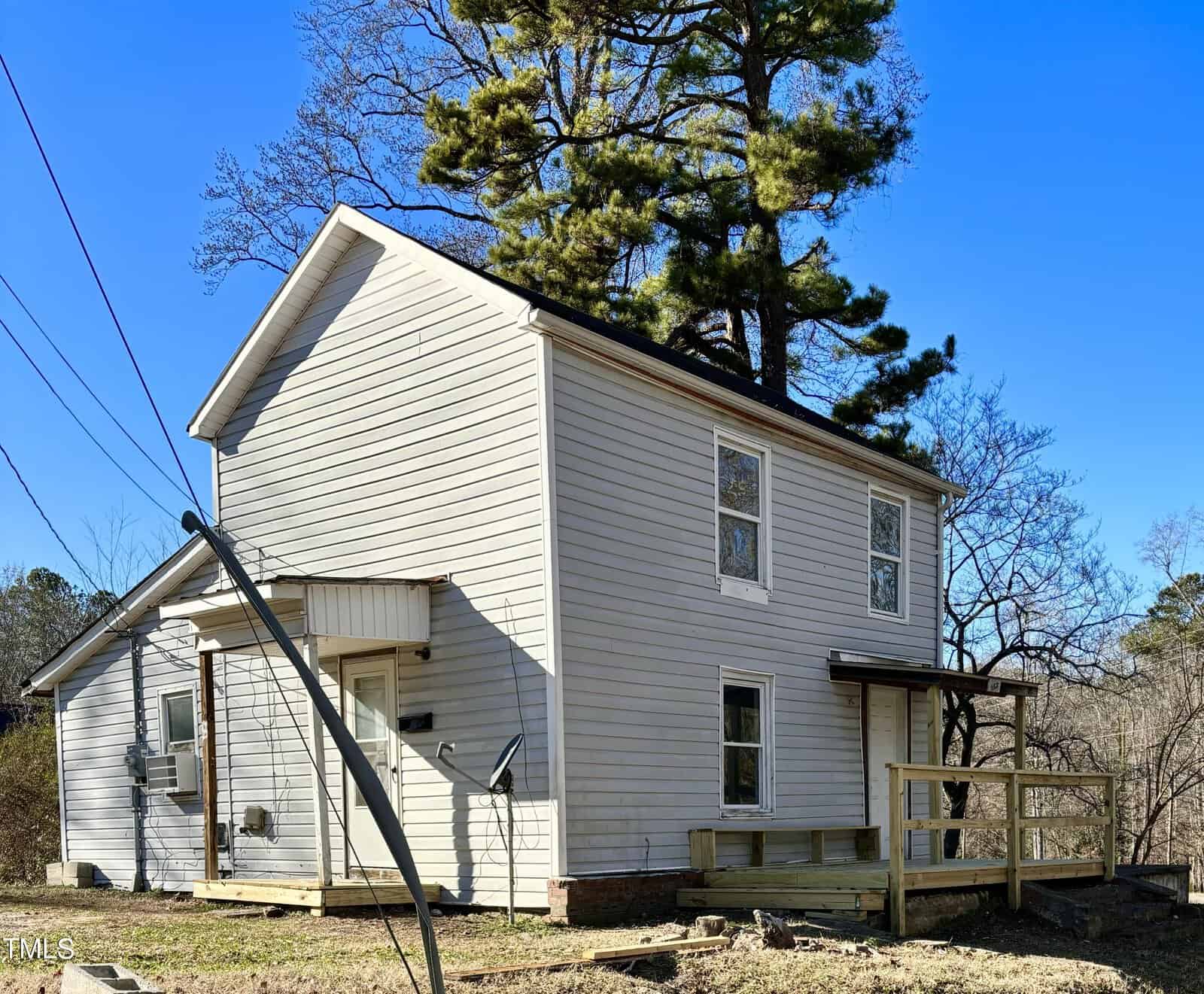 Two-story white house with a side porch and double-hung windows, located on a sloping lot with a backdrop of trees under a clear blue sky.
