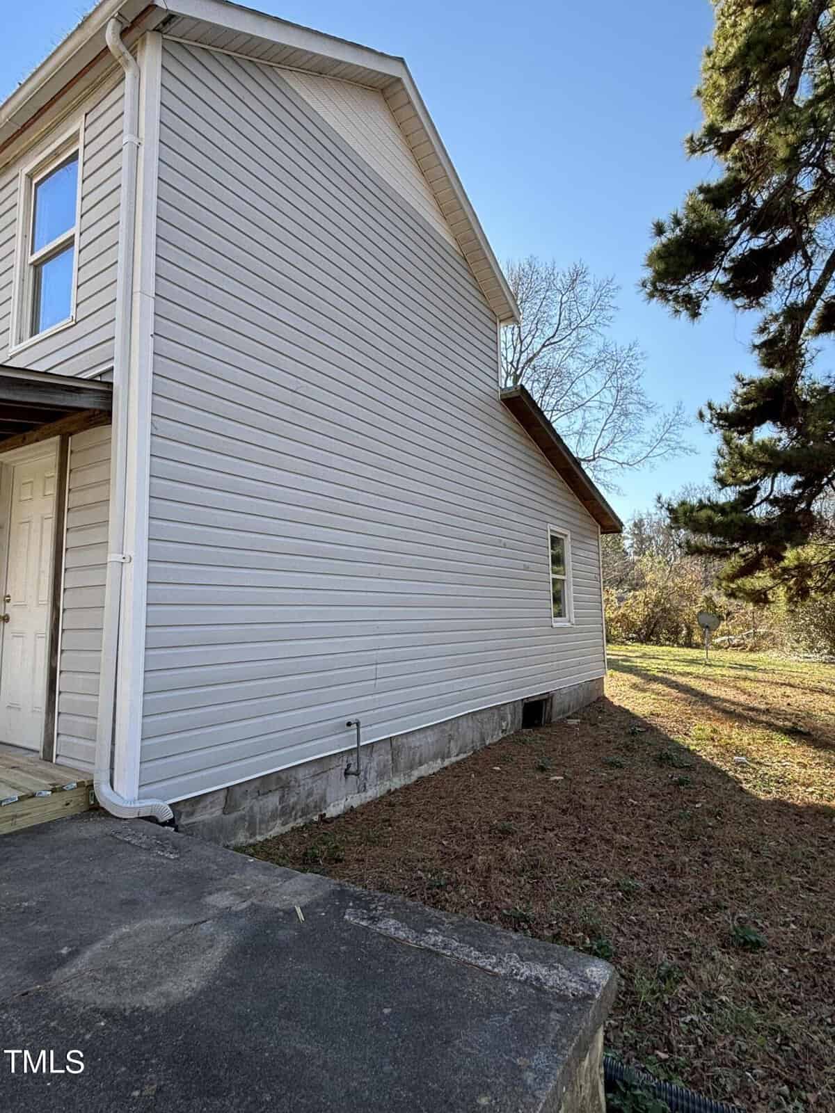 Side view of a two-story white house with horizontal siding, featuring a small window and a concrete foundation. the background shows a blue sky and trees.