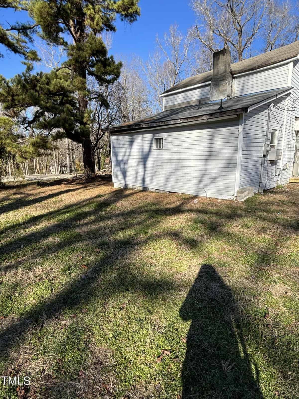 A sunlit backyard showing the shadow of a person and trees on grass, with the side and back of a white house featuring a chimney.