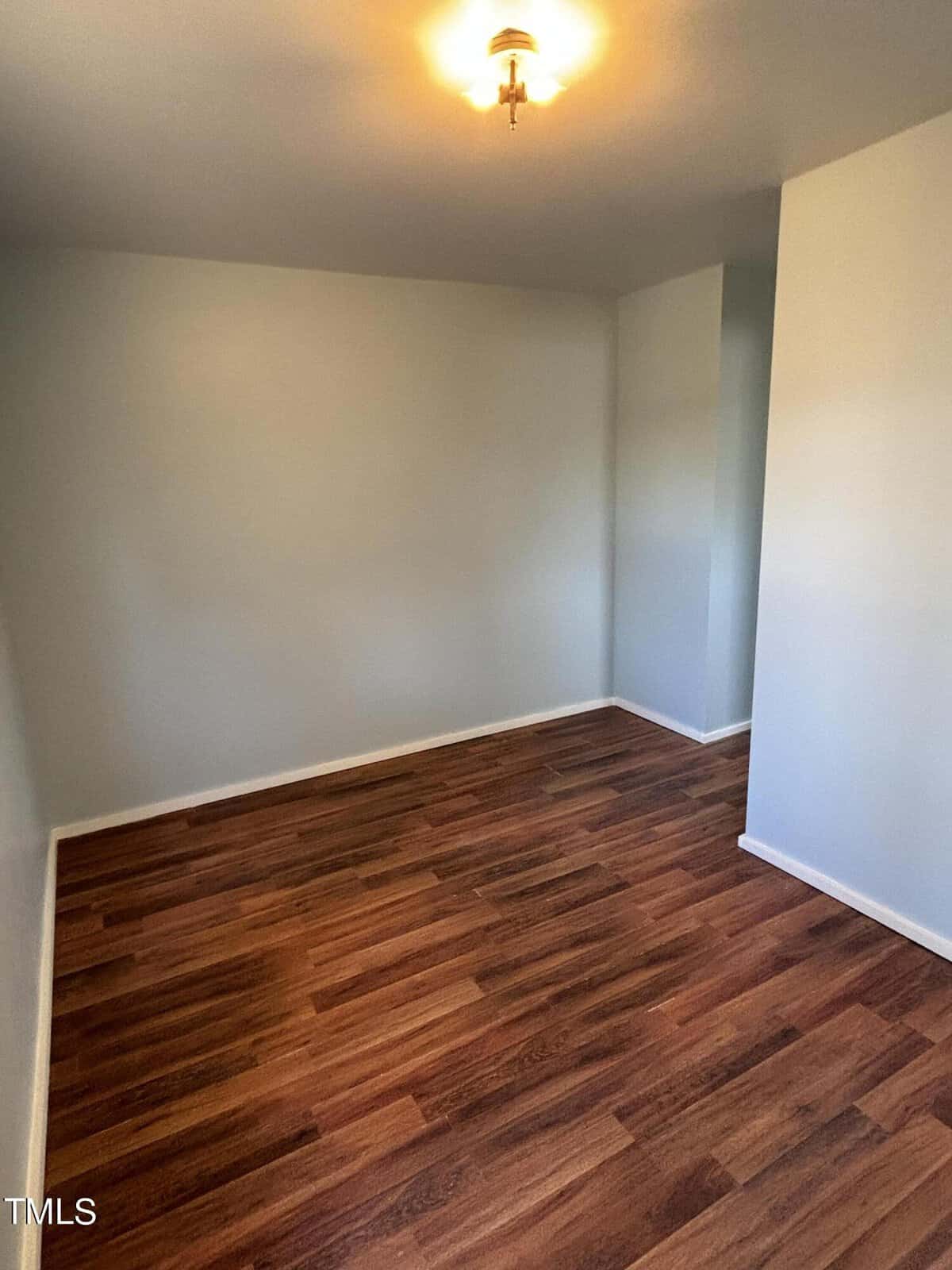 Empty room with wooden flooring, white walls, and a single ceiling light fixture. the corner of the room shows a change in wall angle, presenting a small nook.