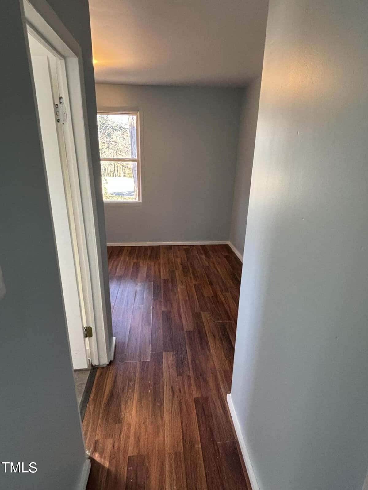 Narrow hallway in a house with light grey walls, dark wooden flooring, and a white door slightly ajar on the left. a sunlit window at the end illuminates the space.