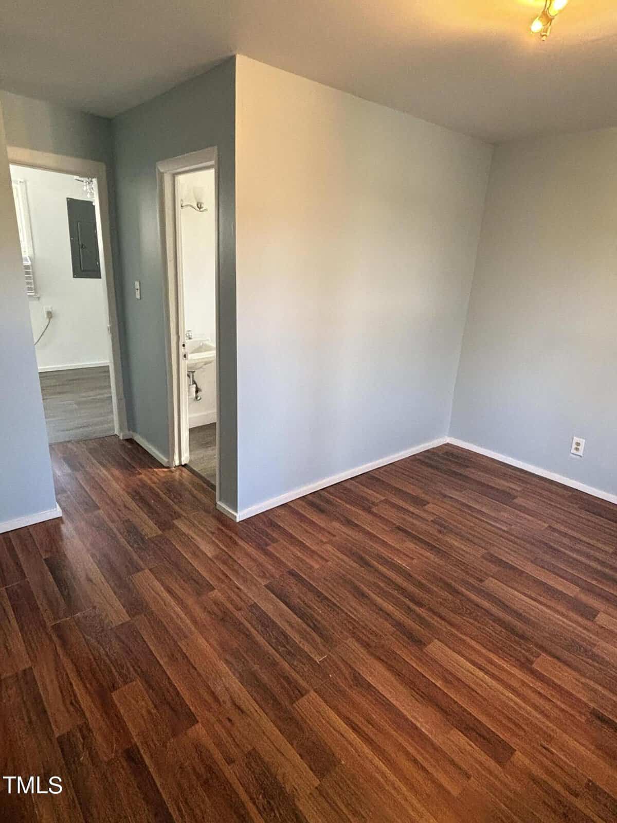 An empty room with polished wooden flooring, light gray walls, and a white ceiling. an open doorway on the left leads to a bathroom. the photo includes a small watermark labeled "tmls" at the bottom.
