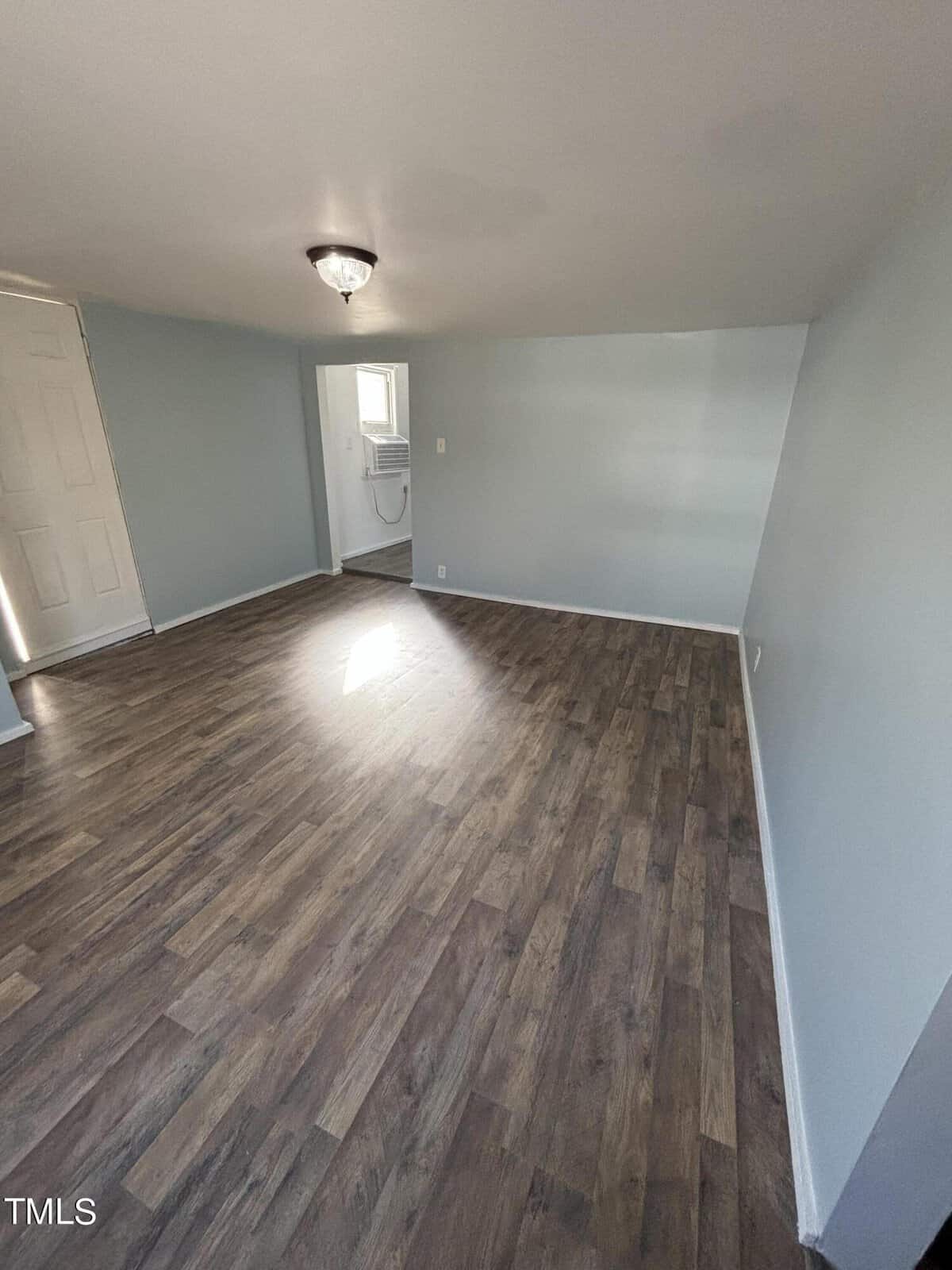 An empty room with light gray walls and dark wood laminate flooring. the room features a single window and a closed white door, with sunlight casting a bright spot on the floor.