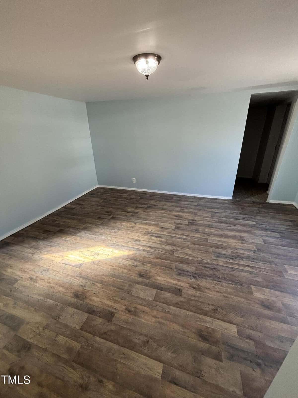 Empty room with light blue walls and dark wood laminate flooring, featuring a ceiling light fixture casting a glow on the floor near the center. an open doorway is visible in the back corner.