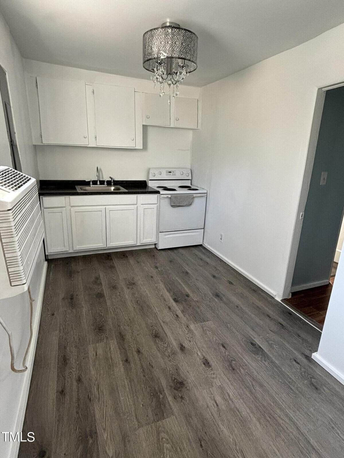 A small kitchen with white cabinets, dark countertops, and white appliances. features gray wood-style flooring and a chic chandelier.